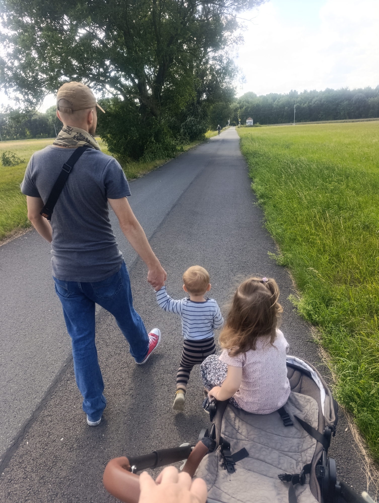 The image depicts a family walking on a paved path through a rural or park-like setting, possibly near Olomouc, Czechia. The foreground shows the back of a man wearing a grey t-shirt, blue jeans, a tan baseball cap, and a patterned neck gaiter. He is holding the hand of a young child, a boy with blonde hair, wearing a striped long-sleeved shirt and dark pants. The boy is walking in front of the man, slightly to the right. To the right of the boy and further ahead is a young girl sitting in a stroller. She has long brown hair and is wearing a light-colored shirt with a pattern. The stroller is a dark color with grey padding and is partially visible. The path appears to be an asphalt surface, bordered on the right by tall, green grass. On the left side of the path, there are trees and foliage. In the background, the path continues into the distance, lined by a dense forest on the right and trees and fields on the left. The sky is bright, indicating daylight, with some clouds visible. The perspective suggests the photo is taken from someone walking behind the stroller, possibly another family member.