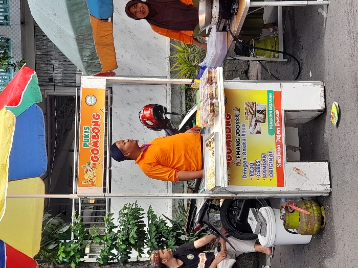 The scene takes place in Kecamatan Sidoarjo, Indonesia, at the coordinates -7.442 latitude, 112.705 longitude. A man wearing a bright orange shirt and a blue cap stands behind a food cart with a sign that reads "Pukis Gombong." He is facing the camera, with his arms crossed. To his left, a man in a green shirt sits on the edge of a sidewalk, looking away from the camera. To the right of the food cart, a woman wearing a hijab is standing, facing away from the camera. She is looking down at the food preparation taking place in front of her. There is a motorcycle parked next to the man sitting on the sidewalk. The food cart is decorated with drawings of fruit and a large sign that lists the types of "Pukis Gombong" offered. The cart is surrounded by various containers of food and preparation equipment. There is a red gas cylinder lying on the ground next to the cart. An umbrella covers the food cart and a second umbrella is open on the right side of the scene. The scene is set in a street environment with a wall and some green shrubs in the background. The pavement is a light gray color. The ground is littered with debris. There are some buildings visible in the background.