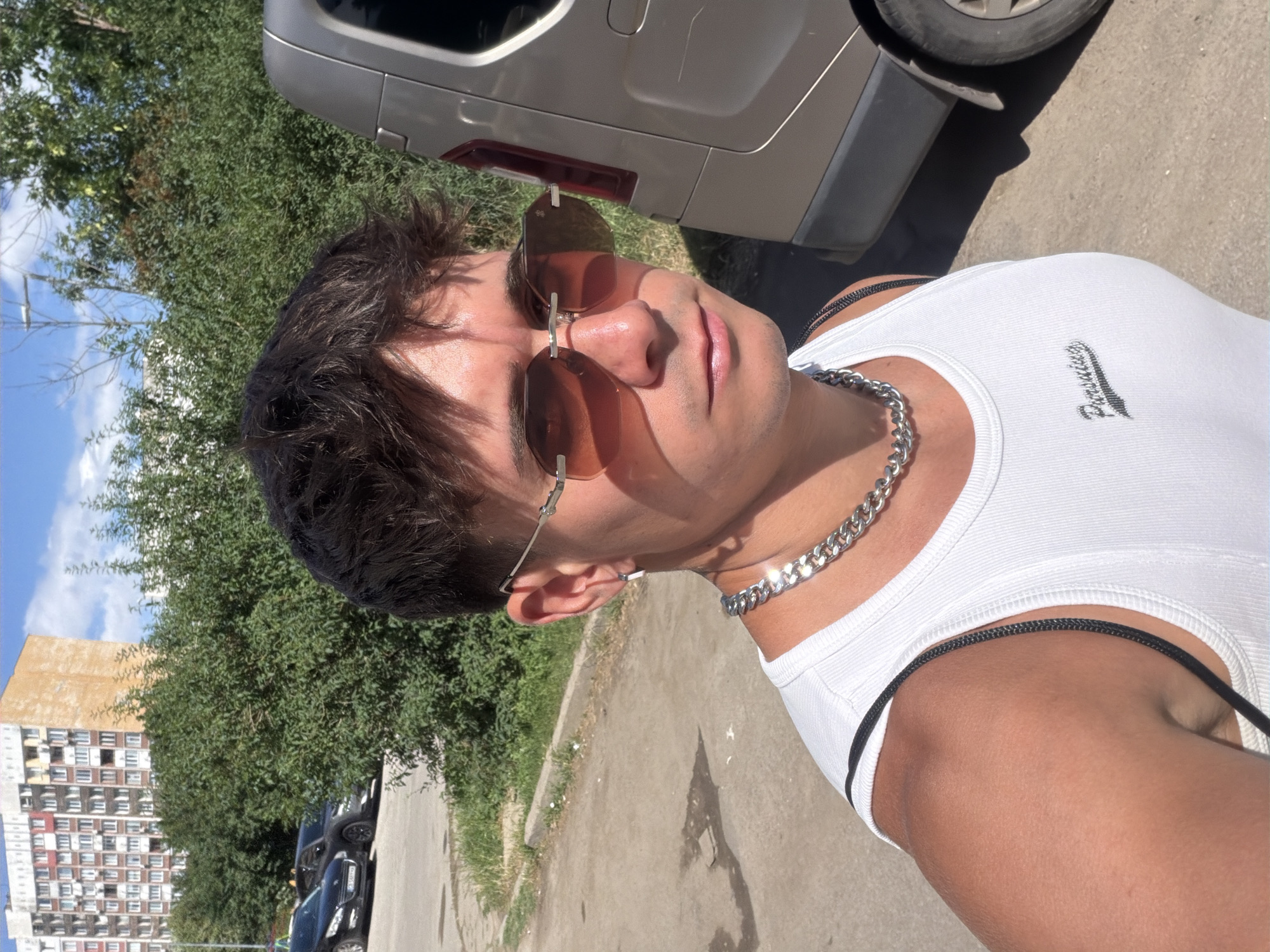 The image shows a young man taking a selfie outdoors on a sunny day. He is wearing a white ribbed tank top and a thick silver chain necklace. He has on brown-tinted square sunglasses. His dark hair is styled messily. The background includes a silver vehicle, lush green trees, and a long, multi-story apartment building with many windows, characteristic of Soviet-era architecture. The ground is paved with asphalt and some grass is visible. The lighting suggests it is daytime, likely late morning or afternoon, given the strong sunlight and shadows. The man's facial expression is neutral, and the overall mood is casual and relaxed. The location is identified as Sofia, Bulgaria, and the architecture of the building further supports this. No visible text is present in the image.