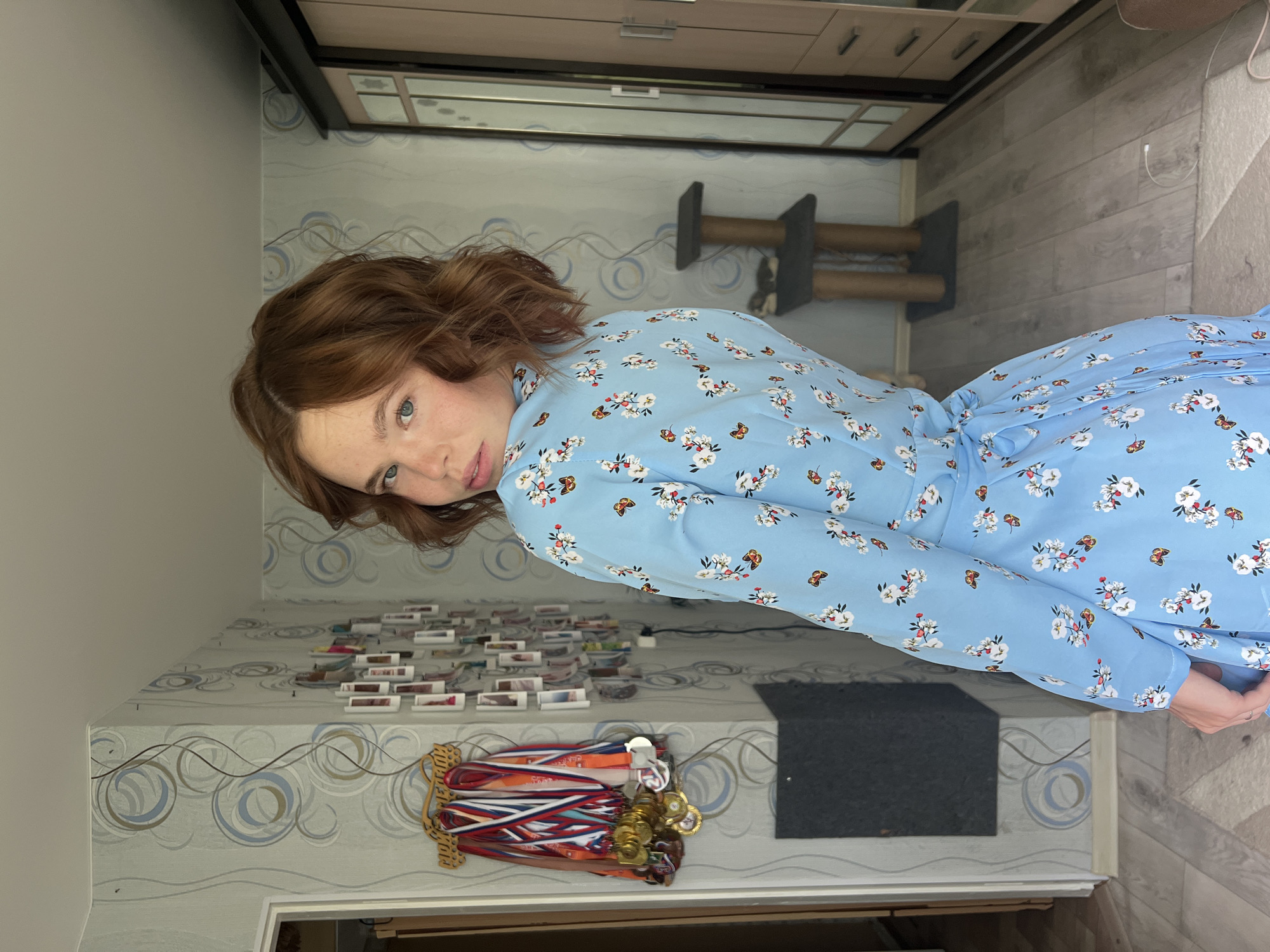 The image depicts a young woman with auburn hair, freckles, and blue eyes, wearing a light blue floral print dress. She is standing indoors, facing away from the camera and looking over her left shoulder. The background shows a room with light blue wallpaper featuring a swirling circular pattern. To the left, a doorway leads to another area, and on the wall, a collection of medals and ribbons is displayed on a wooden rack. Further along the wall are numerous small framed photographs. In the background to the right, a tall wardrobe with mirrored doors is visible, and next to it, a cat tree with two levels. The floor is covered with light-colored laminate flooring. The scene is likely a residential interior. There is no specific information to place this location within the city of Chaikovsky, Russia.