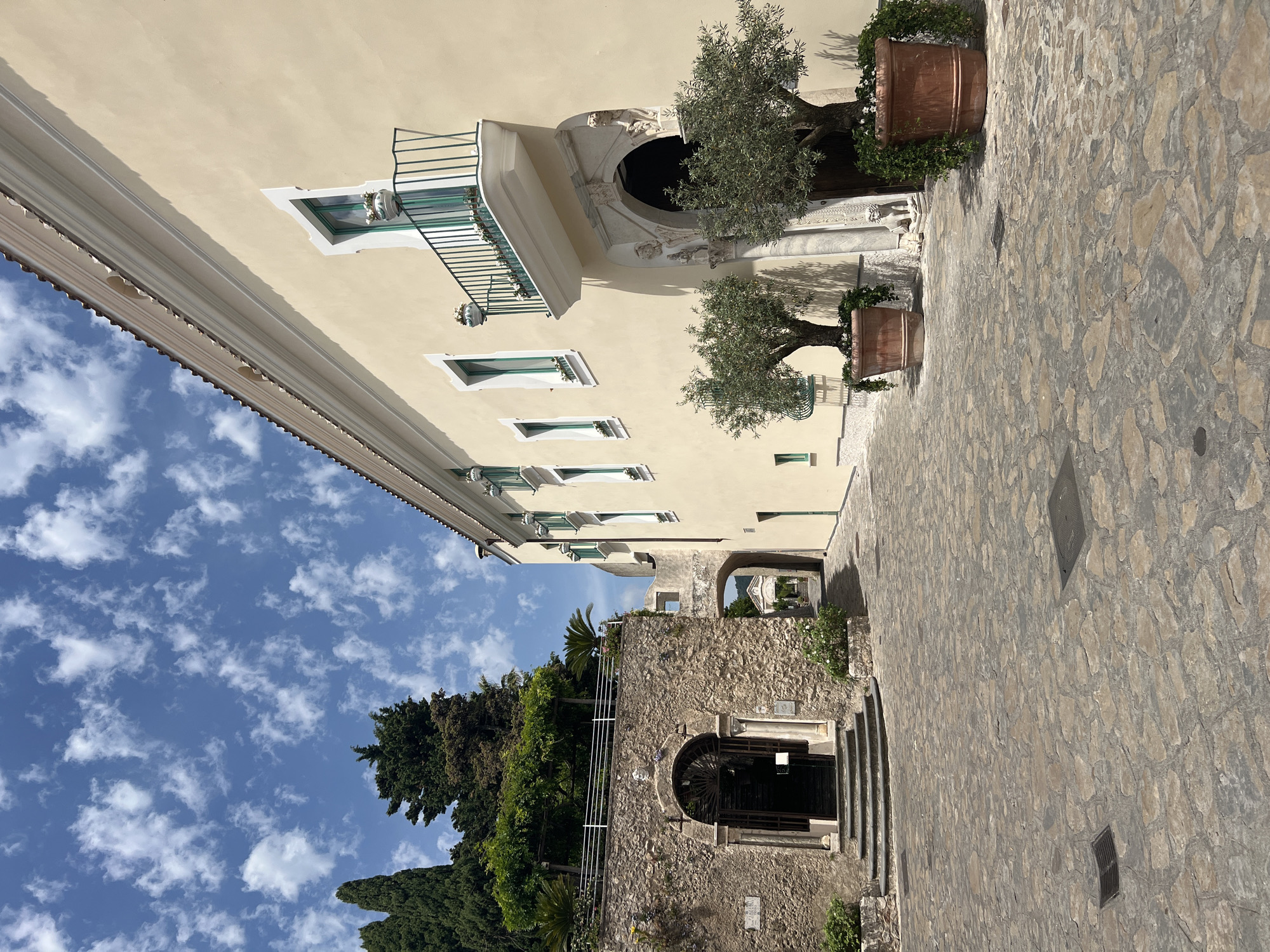 The image depicts a sunlit courtyard in Ravello, Italy, paved with irregular stone slabs. A pale yellow building with green-trimmed windows and a balcony occupies the right side of the frame. Two large potted olive trees flank the entrance to this building, with lush green foliage spilling over their terracotta pots.
To the left, a rustic stone wall features an arched entryway with a dark metal gate, accessible by stone steps. Above this arch, a covered walkway with greenery extends, leading to another arched passage further into the distance. Beyond this second arch, a glimpse of another building and more distant greenery can be seen. Cypress trees and other dense foliage are visible on the left, against a bright blue sky dotted with scattered white clouds. The overall scene conveys a sense of historical charm and Mediterranean tranquility, with no people visible.