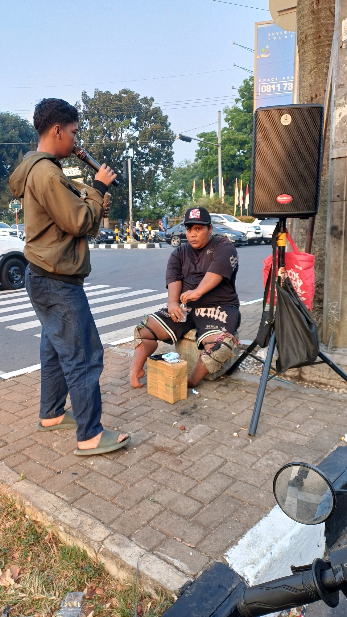 A young male stands on a sidewalk in Cibinong, Indonesia, holding a microphone and facing a seated man. The standing male wears a brown hooded jacket and blue jeans with green slip-on sandals. The seated male, wearing a black t-shirt, black shorts, and a black baseball cap, sits on a stone, holding a bottle of water. A portable speaker is set up to the right of the seated man. A red bag and a black bag hang from the speaker stand. The background shows a street with cars and trees, and a billboard advertising "SPACE AVA 0811 73". The sidewalk is paved with brick-like tiles, and grass lines the edge of the sidewalk.