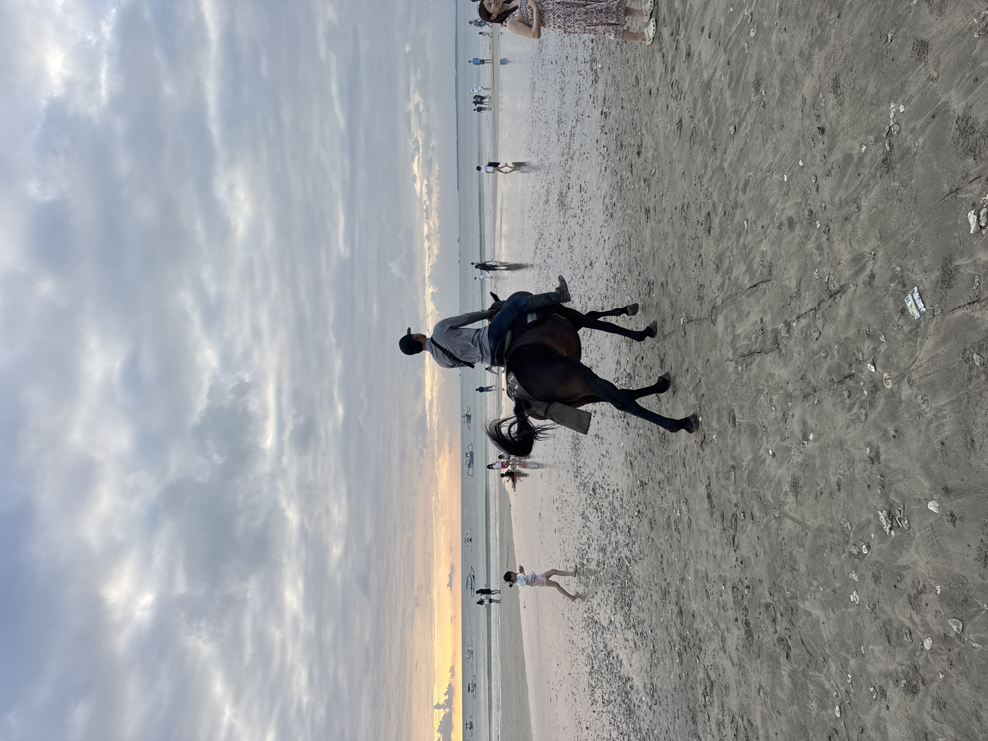 A man rides a dark brown horse along the wet sand of Kuta Selatan beach in Bali, Indonesia, as the sun sets on a cloudy day. The horse is facing away from the camera, towards the ocean, and the rider is wearing a grey long-sleeved shirt, blue jeans, and a black baseball cap. The beach is populated with several other people walking or wading in the shallow water. In the background, numerous traditional fishing boats are visible on the calm sea, and the horizon is marked by a soft orange glow from the setting sun. The sand is scattered with small shells and debris, and the tide has left ripples and pools on the surface. To the right of the horse, a woman in a patterned dress and white clogs stands looking towards the ocean. Further down the beach, a young girl in pink shorts and a white top is walking, also facing away from the camera.