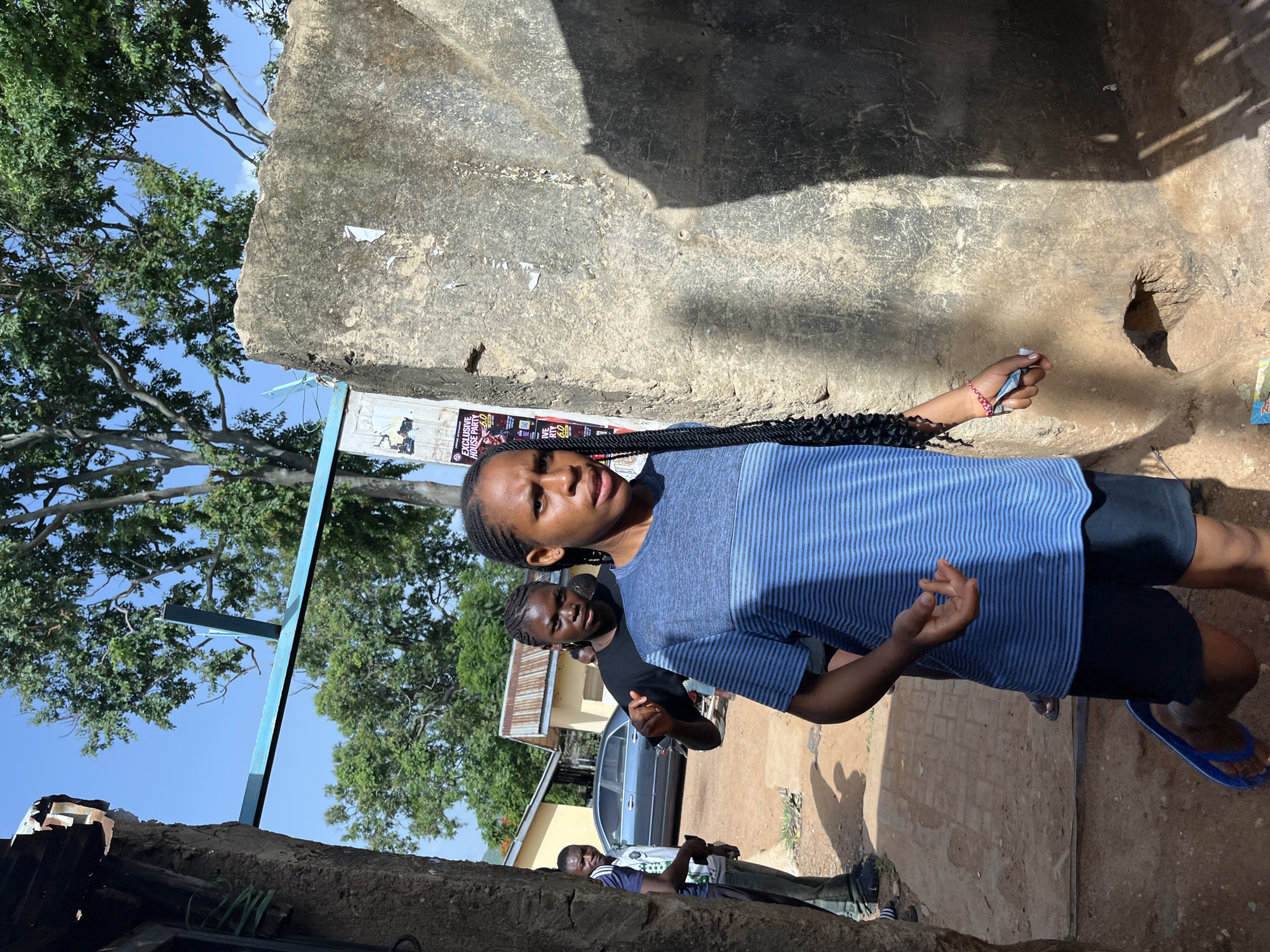 In an outdoor setting, possibly in Jos, Nigeria, a young woman with long, braided hair is walking towards the camera. She is wearing a blue t-shirt with horizontal stripes and dark shorts, along with blue flip-flops. In her right hand, she is holding what appears to be some folded paper or currency. Behind her, another young woman in a black t-shirt is walking and looking towards the camera. Further back, a man in a white and blue striped shirt stands beside a car, with a building behind him. The environment appears to be a street or pathway with rough, textured walls and some vegetation visible in the background. The lighting suggests it is daytime with a bright sun.