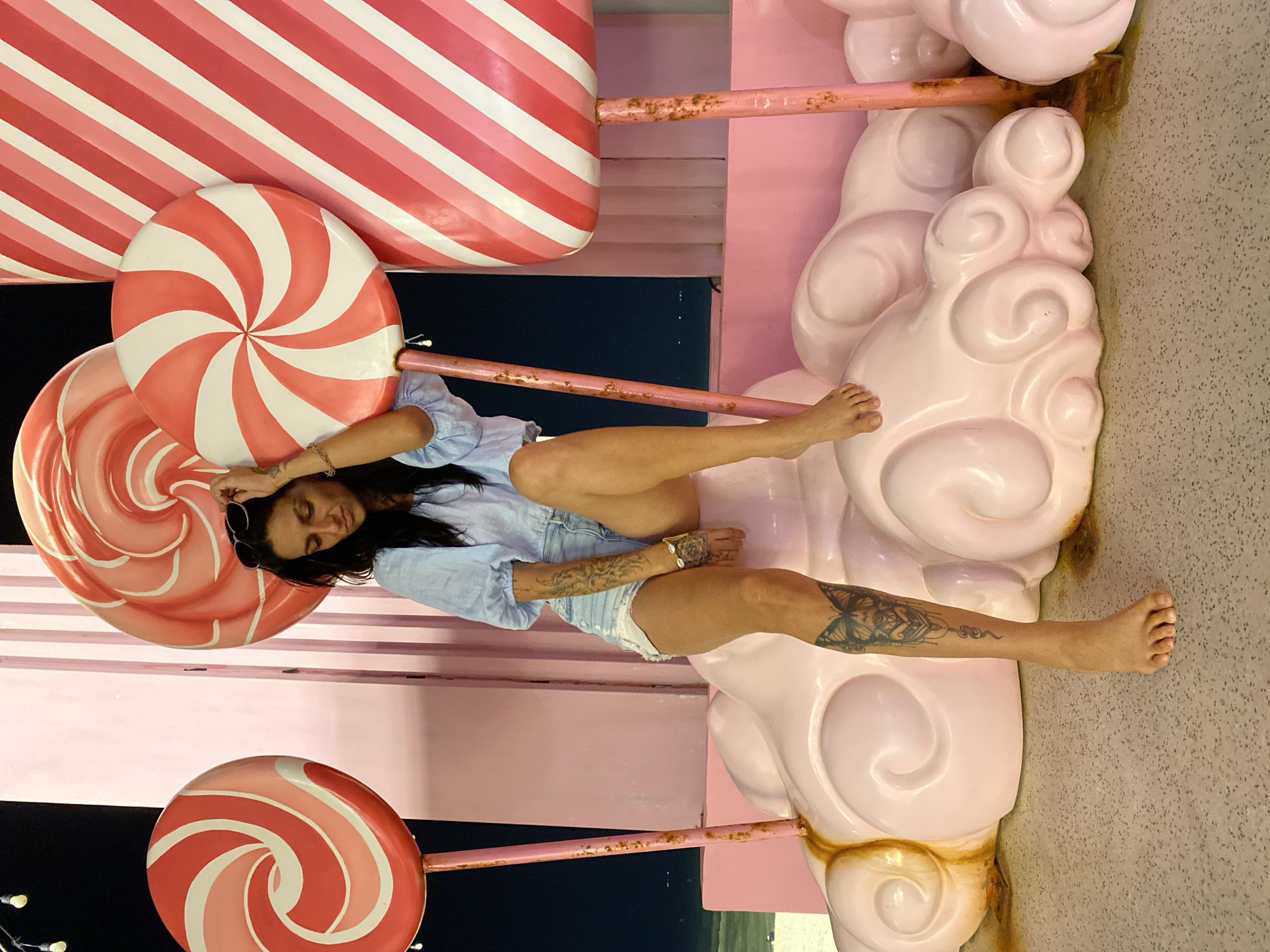 A woman with dark hair is sitting on a pink, cloud-like structure in front of large, colorful lollipops. She is wearing a light blue blouse and denim shorts. Her legs are bare, and she has tattoos on her left leg. She is holding a pair of sunglasses in her right hand and looking down. The background consists of large, striped lollipops in shades of pink and white, and a pink structure with vertical stripes. The scene appears to be an outdoor setting at night, possibly an amusement park or a themed attraction. There are faint lights visible in the distance. The ground is a textured grey surface. The location is identified as Tien Thanh Ward, Vietnam.