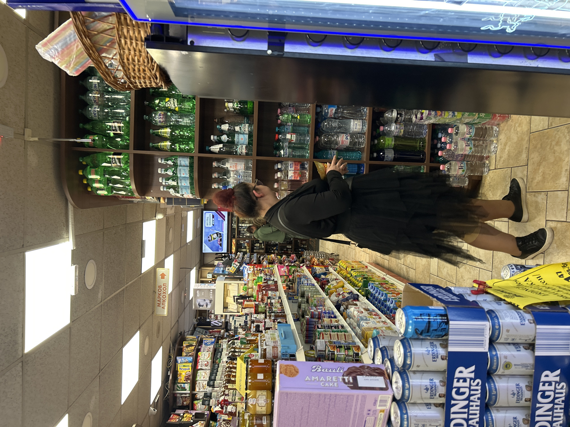 Here is a description of the image:
**Scene:** The photograph was taken inside a well-stocked convenience store. The store is brightly lit with fluorescent ceiling lights, and the tiled floor is a neutral beige color.
**People:** The main subject is a person standing near the beverage section. They have their back to the camera and are looking at the shelves of bottled water. They are wearing a black dress with a tulle skirt, a dark jacket, and black shoes.
**Objects:**
* Shelves stocked with various bottled water brands.
* Boxes of Erdinger beer stacked on the floor.
* A box of "Barnii Amaretti Cake" visible on the shelf.
* Signage in Russian and other languages (likely Cyrillic) is present, indicating the direction of alcohol products.
* A vending machine is present above the bottled waters.
* A basket is seen near the ceiling.
**Actions and Interactions:** The person is browsing the selection of bottled water.
**Environment:**
The environment appears to be a typical convenience store, with shelves packed with a variety of goods, ranging from snacks and drinks to household items. The presence of Cyrillic signage suggests the store is located in a region where this language is prevalent.
