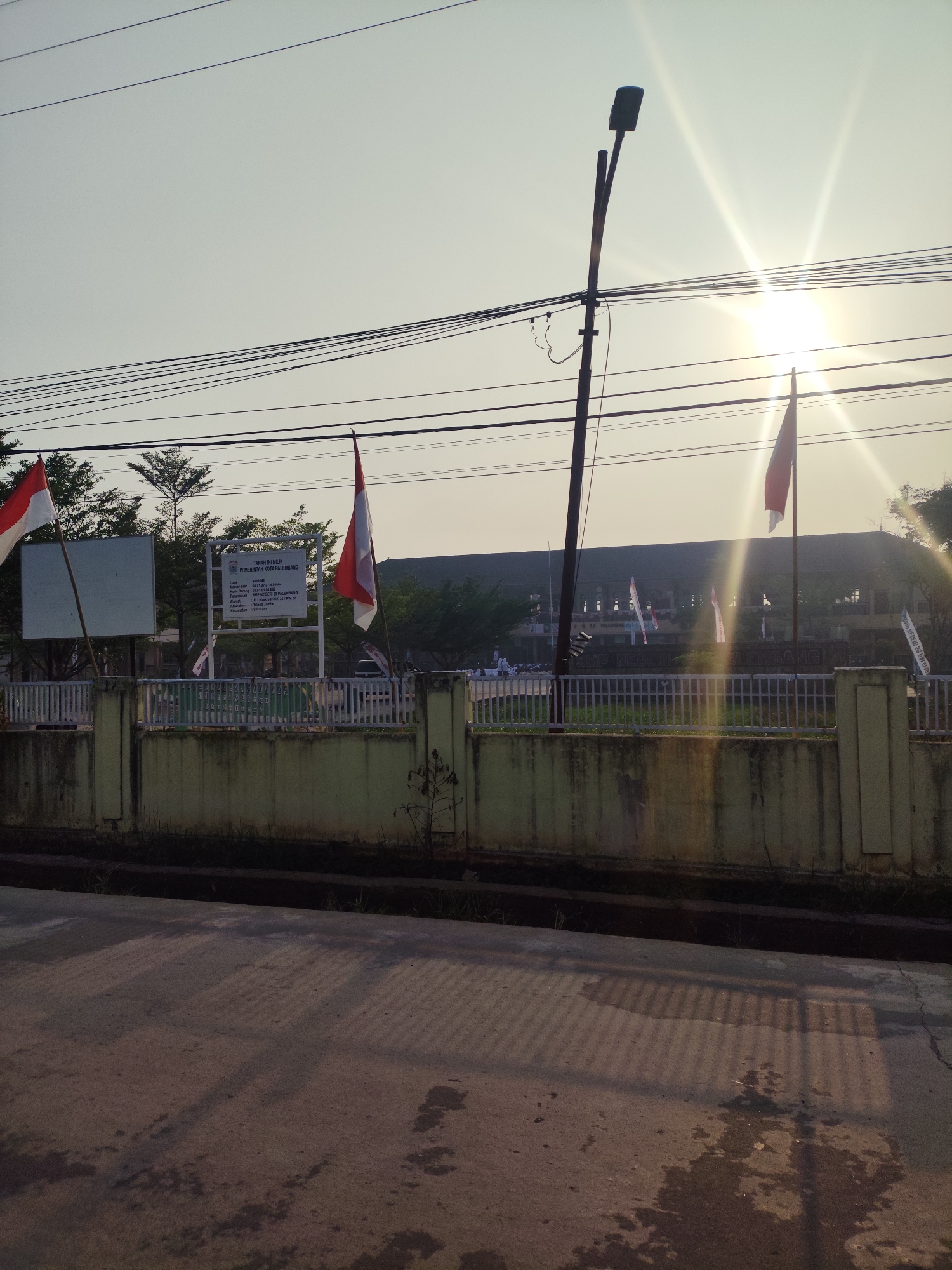This image depicts the exterior of SMP Negeri 16 Palembang, a public junior high school located in Palembang, Indonesia. The sun is low in the sky, casting a bright glare and long shadows across the scene, suggesting either sunrise or sunset. Two Indonesian flags are prominently displayed, one to the left of a white informational sign and another to the right. The sign itself contains text in Indonesian, indicating it is government property and likely provides information about the school or its land. The school building, a long, dark structure, is visible in the background behind a white metal fence. The foreground shows a concrete sidewalk and road. Overhead, a network of power lines crisscrosses the sky, and a street lamp stands near the center. The overall atmosphere is one of a typical school day or an event taking place outdoors.
