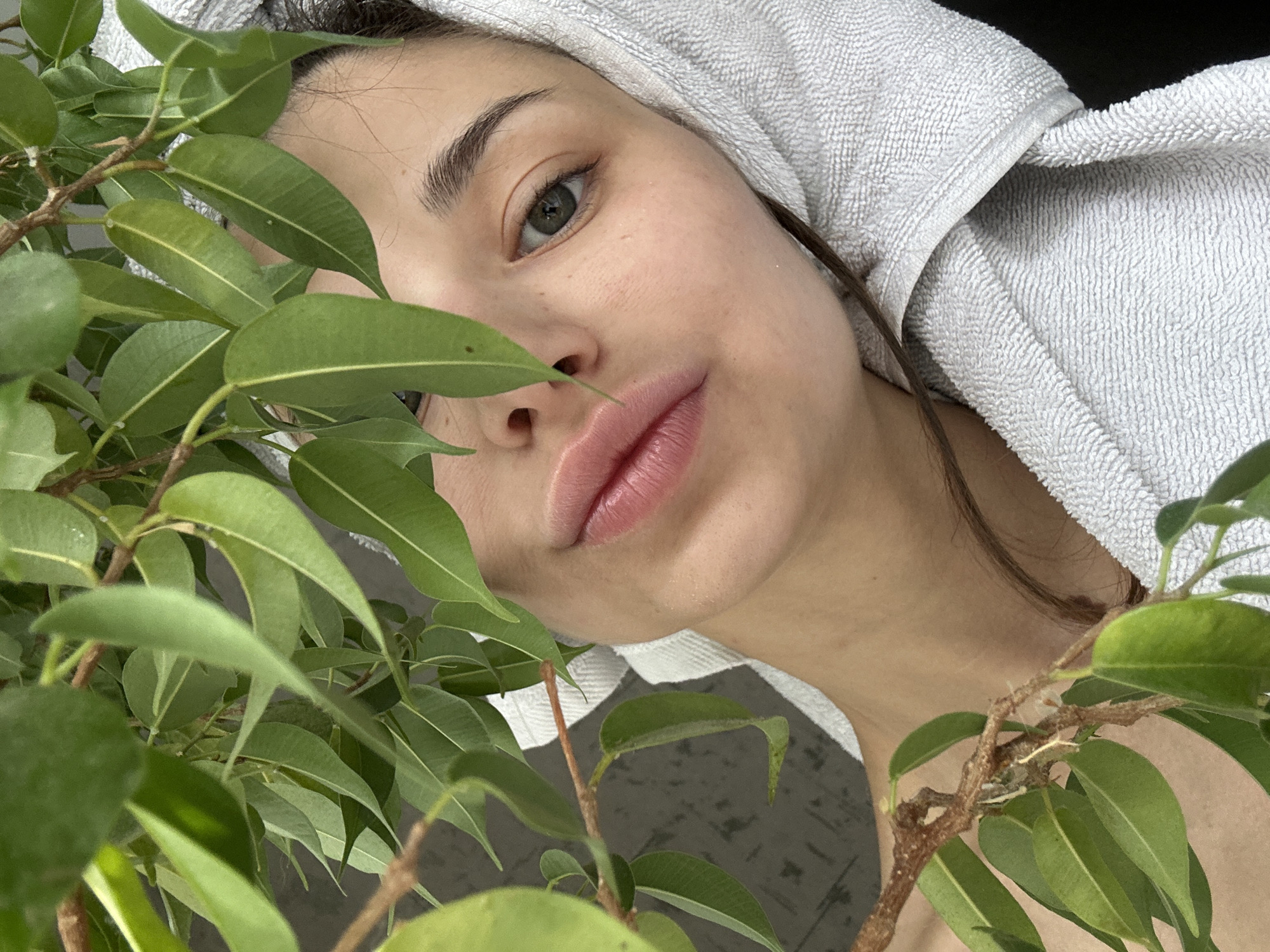 A close-up photograph shows a person with fair skin and brown hair partially obscured by green leaves from a plant. The person is wearing a white towel wrapped around their head, with strands of brown hair visible. The person has light eyebrows, one visible eye with a grey iris, and full lips. The plant occupies a significant portion of the left side of the frame, with some branches extending into the right side. The background is indistinct, but appears to be an interior space. There is no evidence to suggest the photograph was taken in Izhevsk, Russia, or to provide any forensic details. The image is a portrait of a person with a plant.