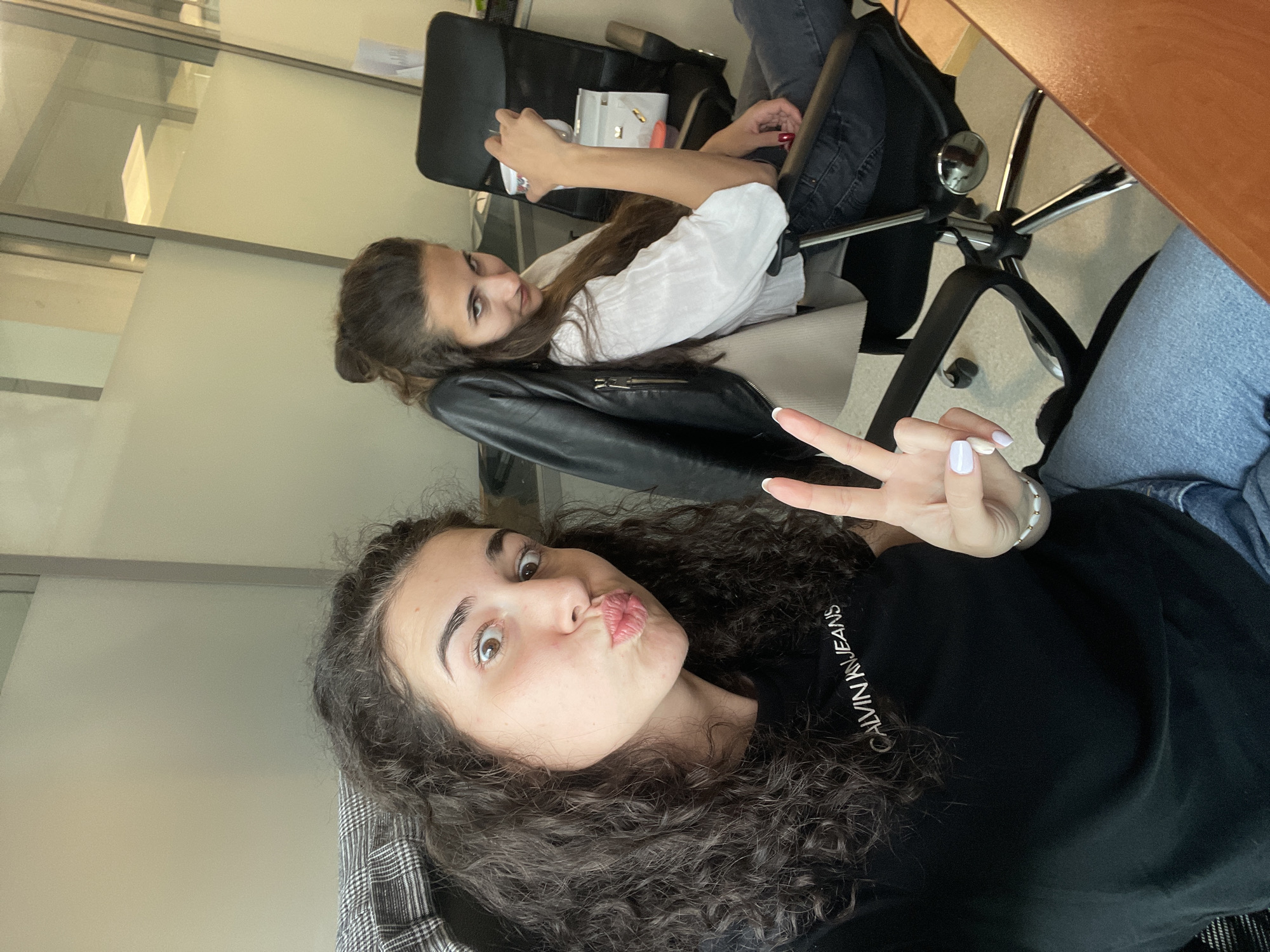 Two young women are in an indoor office setting. The woman in the foreground has curly brown hair and is wearing a black Calvin Klein t-shirt. She is pouting her lips and giving a peace sign. The other woman is in the background, sitting in a black office chair, and wearing a white shirt with a black leather jacket over it. The office has a light-colored wall with glass partitions. A wooden table is visible in the upper right corner. This is likely in Sofia, Bulgaria, due to request to include the location in the city of Sofia, Bulgaria.