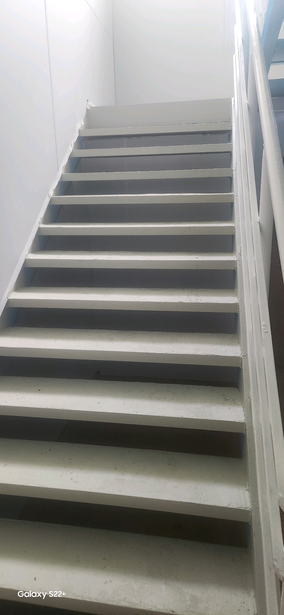 This is a photograph of an indoor staircase, likely in a utilitarian building such as a stairwell or fire escape. The stairs are constructed of concrete and appear to be painted white, with some signs of wear and tear on the paint. The risers are open, creating a series of horizontal planes. A white railing is visible along the right side of the staircase. The surrounding walls are also painted white, and the lighting suggests an overhead source, casting shadows beneath each step. The overall impression is one of simplicity and functionality. There are no people present in the image. The location in Nyurba, Russia, is not visually evident from the photograph itself; this information is provided externally.