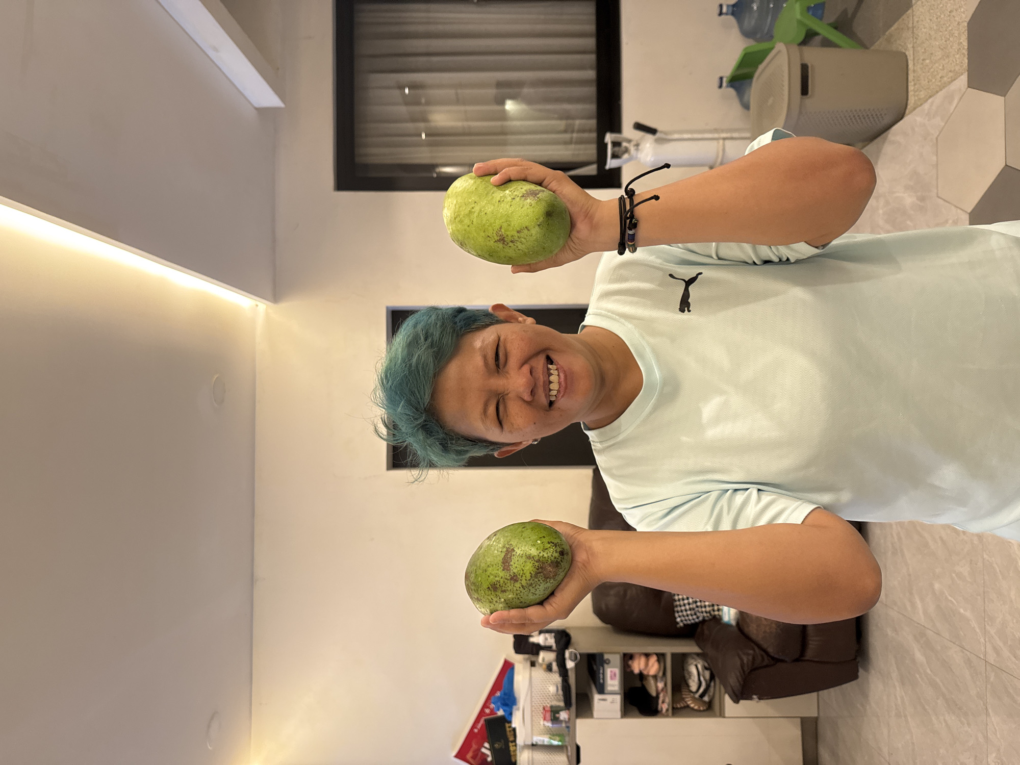 A person with short, dyed blue hair and a light blue t-shirt stands indoors, holding two large green fruits, possibly avocados or mangoes, one in each hand. They are smiling broadly, looking directly at the camera. The background includes a window with blinds, a brown leather armchair, a white cabinet with items on shelves, and a gray trash can with a water bottle on top. The floor is tiled with a geometric pattern. The location is implied to be Bekasi, Indonesia.