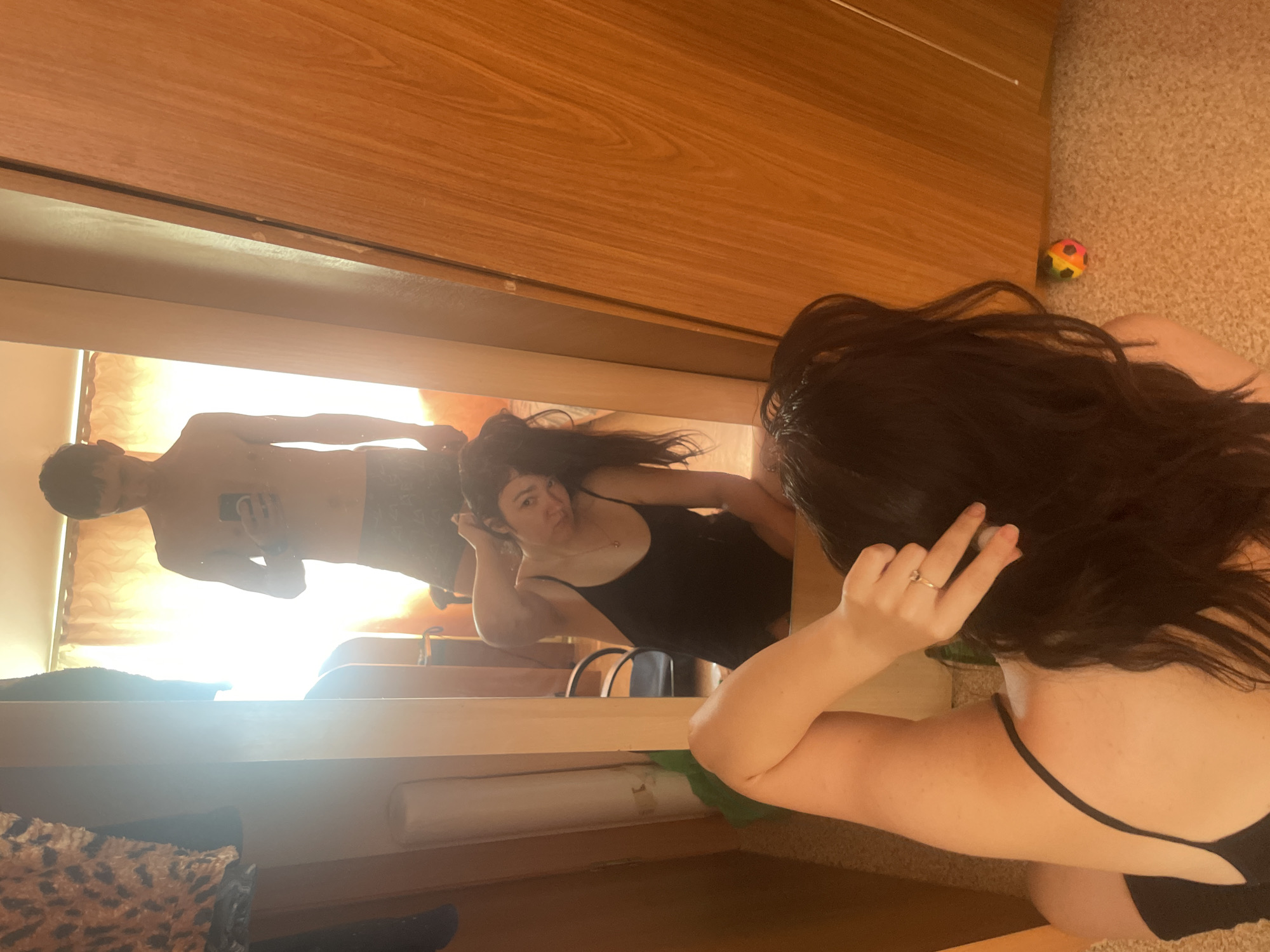In a room with wood-paneled walls and a carpeted floor, a young woman with long, dark hair is taking a mirror selfie. She is wearing a black bikini top. Her reflection shows a young man, shirtless and wearing patterned shorts, also taking a selfie in the mirror. The setting appears to be indoors, possibly a bedroom or a dressing area, with curtains visible in the background of the reflection. A small, colorful ball toy is on the floor to the right of the frame. The lighting suggests it could be daytime, with natural light filtering in from behind the curtains. The woman's expression in the reflection seems playful or quizzical. There is no visible text in the image.