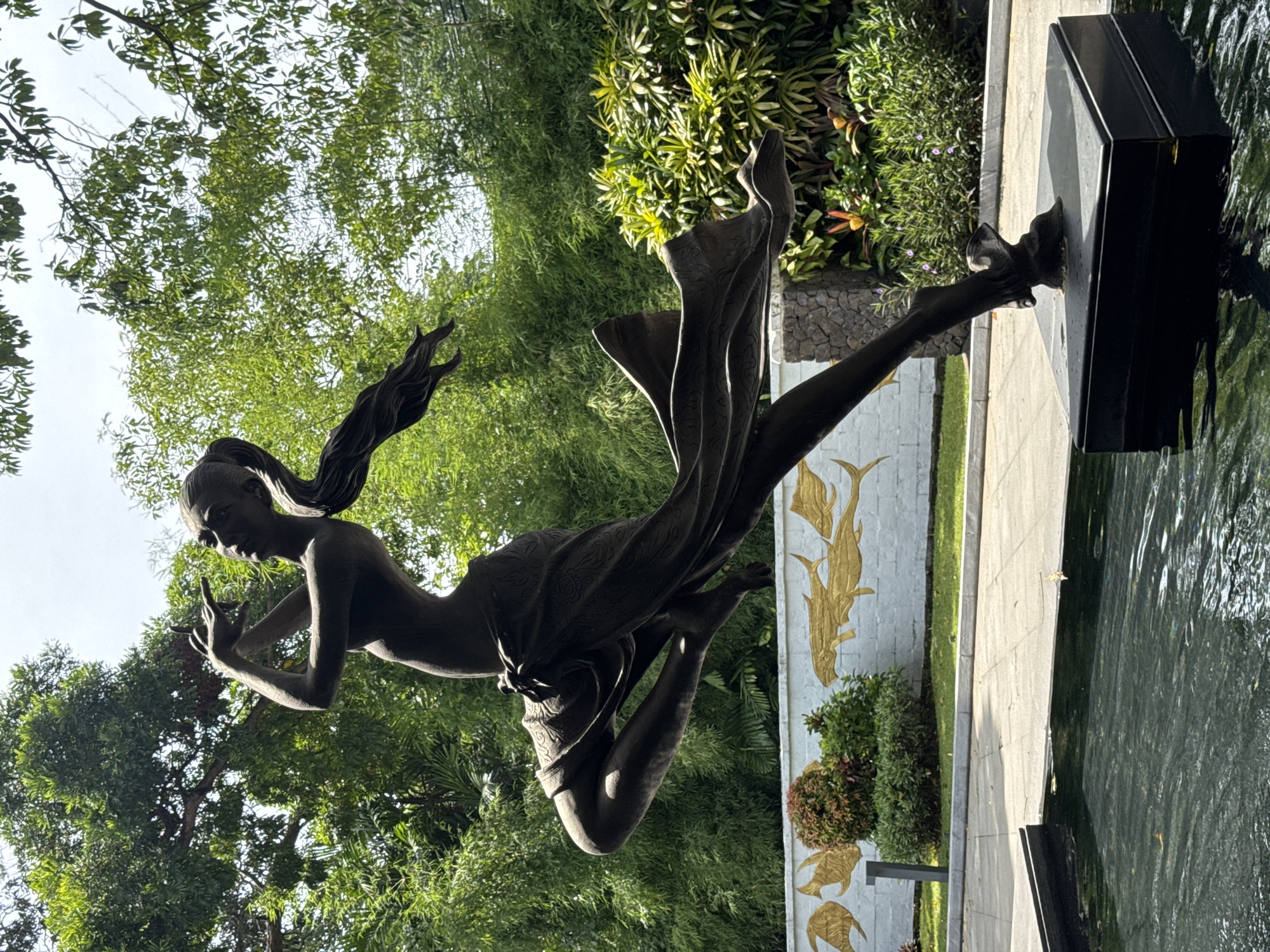 The image shows a bronze statue of a woman in motion, possibly dancing or flying. She has long hair flowing behind her and is partially draped in fabric. The statue is positioned near a water feature, possibly a pond or pool. The lower part of one leg is connected to the water feature using a black rectangular structure.
The statue is set against a backdrop of lush green vegetation, including trees and bushes. There's a wall in the background adorned with golden fish-like figures. The environment suggests an outdoor setting, possibly a garden or park.
The image was taken in Kuta Selatan, Indonesia.