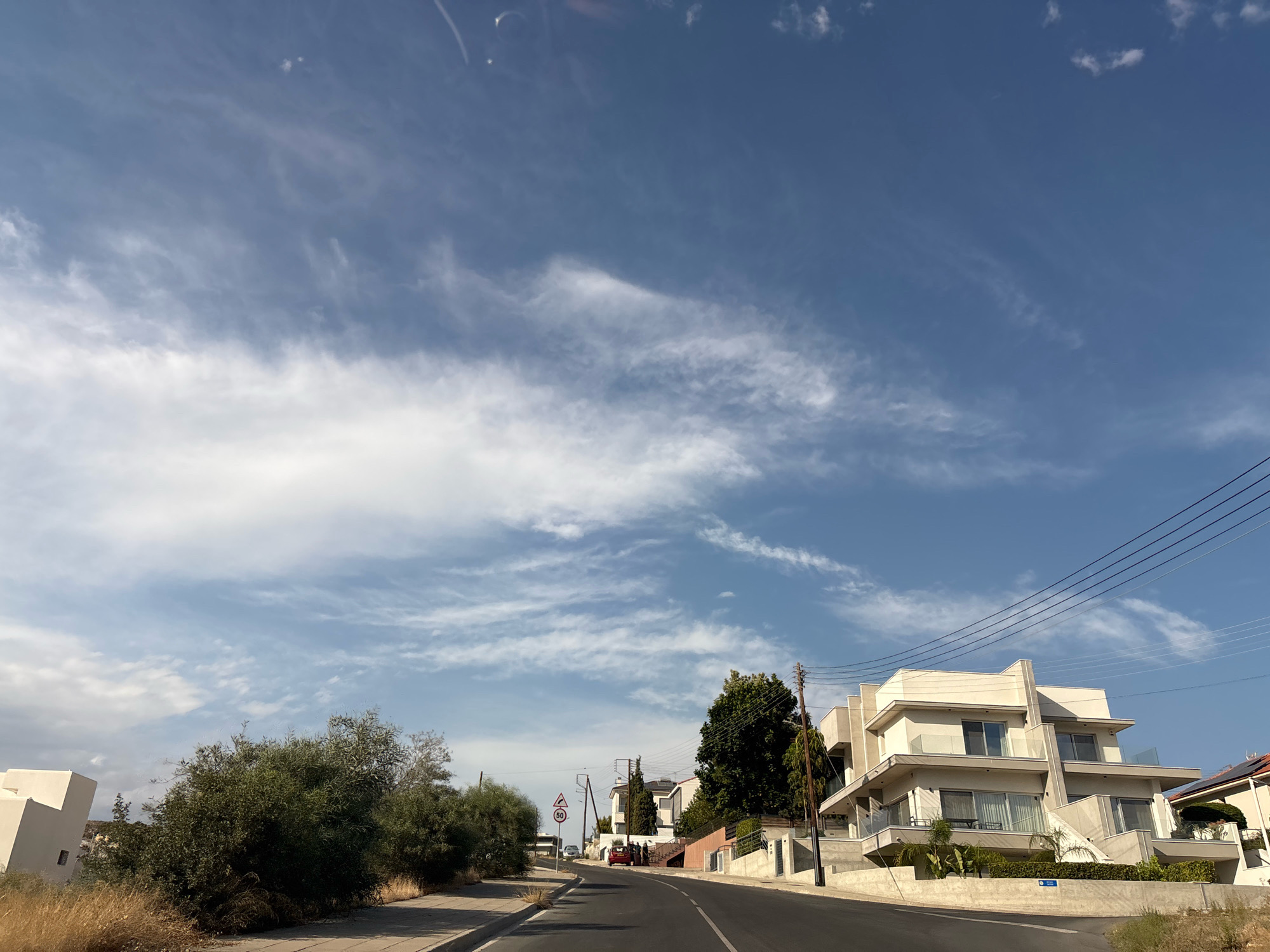 This image depicts a street scene in Agios Athanasios Municipality, Cyprus, under a partly cloudy blue sky. The road curves to the right, with a sidewalk on the left and a row of houses on the right. Several modern, multi-story houses with white and beige facades and glass balconies line the right side of the road. Lush green trees and vegetation are visible, with some dry, tall grass along the sidewalk. There are power lines running overhead supported by utility poles. A red car is parked on the side of the road, and a few people are gathered near it. Traffic signs, including a speed limit sign (50), are present. The overall impression is a residential area during daylight.