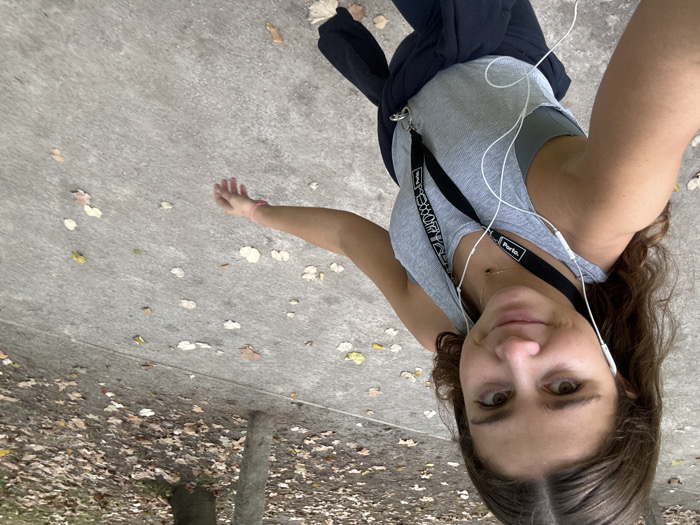A young adult female is depicted in a selfie-style photograph taken outdoors on a paved pathway in Aldoar, Foz do Douro e Nevogilde, Portugal. She has brown, wavy hair and is looking directly at the camera with a slight smile. She is wearing a light grey ribbed tank top over a darker grey sports bra or top. A dark blue or black jacket is tied around her waist. White wired earbuds are inserted into her ears, with the wires extending downwards. Two black lanyards are visible around her neck; one has "Porto." written in white. A delicate necklace with a dark pendant is worn around her neck. A light pink band is present on her right wrist. The pathway is a light grey, textured concrete surface with scattered dry, brown, yellow, and beige leaves. To the right and in the background, a wooded or park-like area with tree trunks and a denser covering of fallen leaves is visible. The scene is illuminated by natural daylight.
