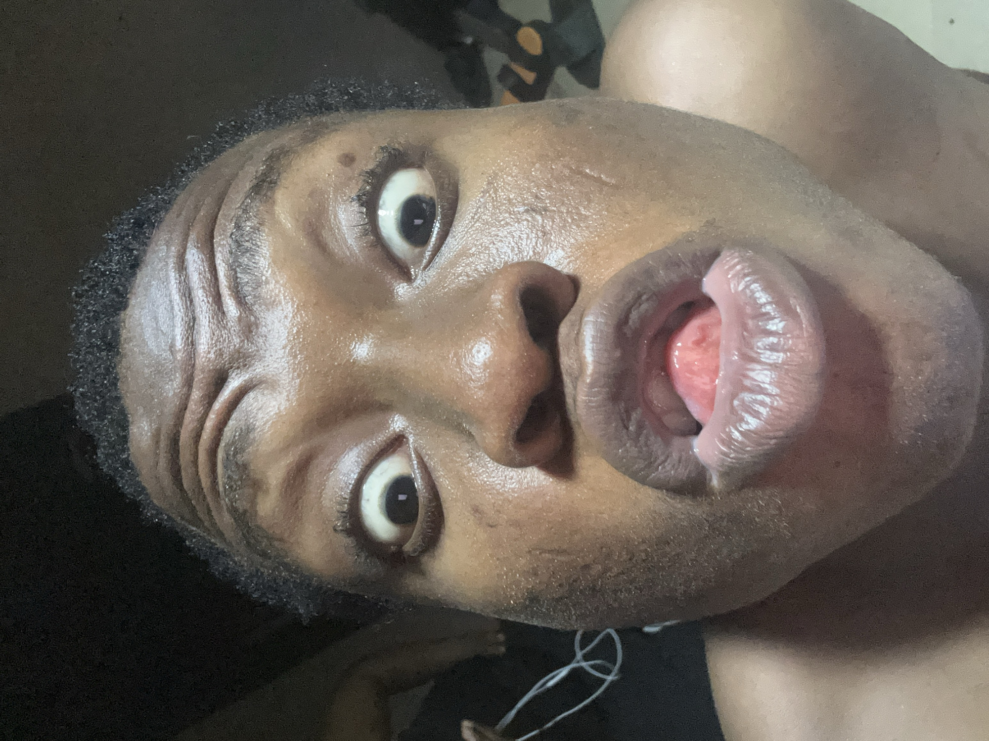 A close-up, distorted, upward-angled shot features a person with dark skin, wide eyes, and an open mouth with the tongue visible. The person's face appears moist or sweaty. The individual has short, dark hair. A white earphone cord is visible near the lower part of the frame. The background is dark and indistinct. No specific location is discernable.