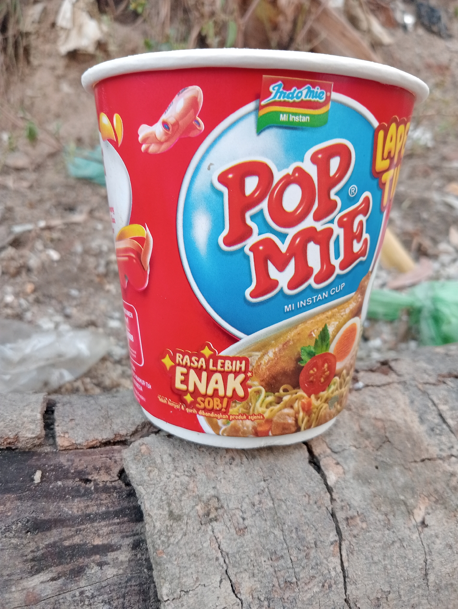 A close-up, eye-level shot shows a red and white cup of Pop Mie instant noodles placed on a rough-hewn, weathered log. The cup is adorned with vibrant branding, featuring the Indomie logo and the Pop Mie name in bold red lettering. A stylized illustration of the noodle dish, complete with an egg and tomato slice, is visible on the front of the cup. In the background, out-of-focus elements suggest an outdoor environment with dirt, debris, and some greenery. The log itself shows significant cracks and textured grain. The setting does not provide specific indicators to pinpoint the location within Kota Negara Timur, Indonesia, beyond it being an outdoor area.