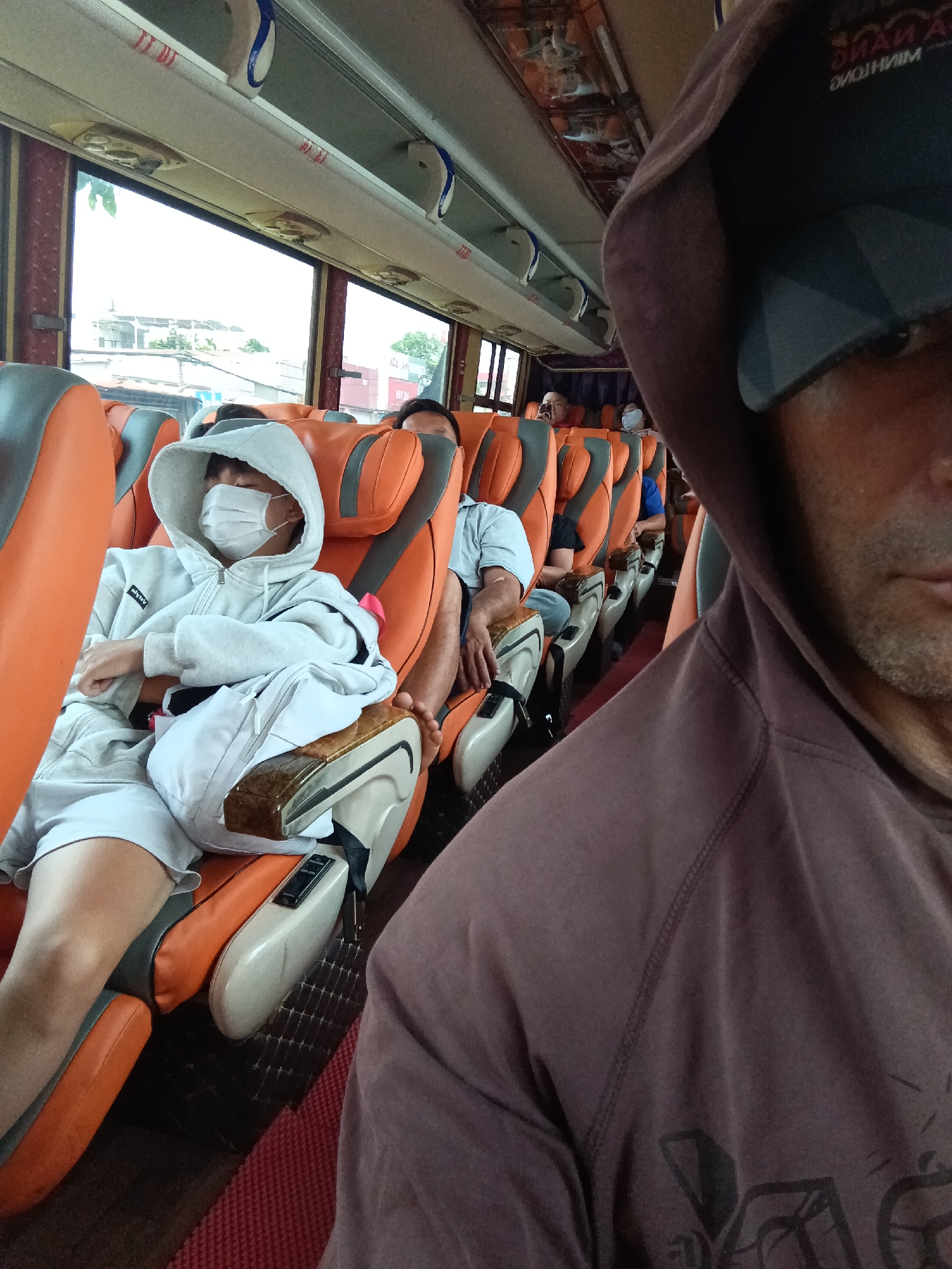 The image shows the interior of a bus with orange and gray seats. In the foreground, a person wearing a brown hooded sweatshirt is partially visible, looking towards the camera. In the middle ground, a person wearing a light gray hooded sweatshirt and a white face mask is seated, appearing to be asleep. This person is wearing a white t-shirt and white shorts. Next to them, another person is seated with their arms crossed. In the background, more passengers are visible on the bus, and through the windows, buildings and trees can be seen. The environment appears to be a public transportation setting.