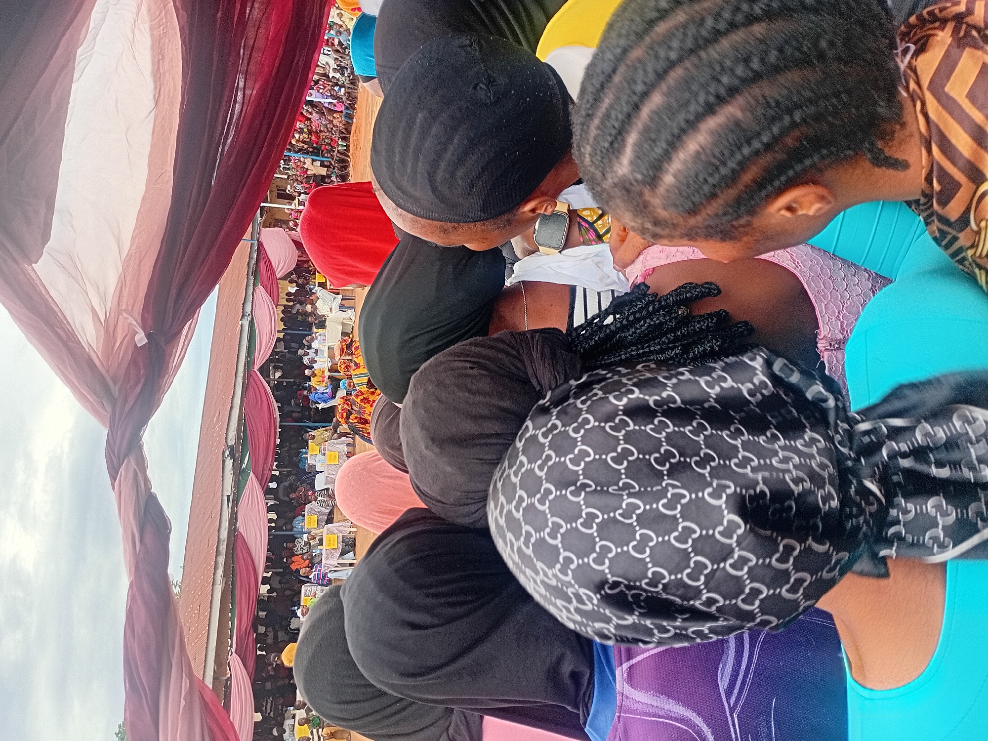The image depicts an outdoor gathering in the city of Ogbogbo, Nigeria, observed from behind a group of attendees in the foreground. The sky is overcast with a light gray and white cloud cover.
In the immediate foreground, the backs and heads of several individuals are visible. Most wear head coverings; specifically, three individuals wear solid black hijabs, one wears a solid pink head covering, and another wears a dark brown head covering. A prominent black headscarf with a repeating pattern of white interlocking 'C' or 'G' shapes is centered in the foreground. Visible clothing includes a bright blue garment, a purple garment with white design elements, a black and white striped top, a pink lace top, and a brown and black patterned top. Braided hairstyles are visible on two individuals on the right side of the frame, one with cornrows and another with longer braids extending from beneath a headscarf on the left. A gold-toned smartwatch is visible on the wrist of an individual to the right. The ground in the foreground and midground appears to be reddish-brown earth.
In the midground, a large group of people is gathered, predominantly seated. Multiple rectangular tables, draped in white fabric, are arranged in rows. Yellow placards or signs are visible on some of these tables. Attendees are seated on chairs around the tables, facing towards an unseen central point or stage. Overhead, large swaths of red and pink fabric are draped, forming a temporary canopy or decorative element for the event.
In the background, a building with a reddish-brown corrugated iron roof is visible behind the crowd and the fabric canopy. Additional people are discernible standing and sitting in denser groups further back. The overall scene suggests a community event, possibly a public meeting, celebration, or competition.