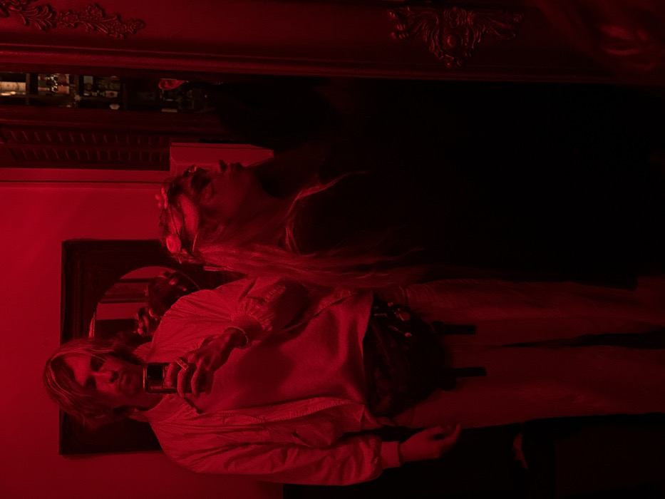 An interior scene is depicted under intense red lighting, creating deep shadows and high contrast. Two individuals are present within what appears to be a dimly lit bar or lounge.
A male subject stands prominently on the left, facing forward. He has light-colored, medium-length hair. He is dressed in a light-colored, collared jacket worn over a lighter-toned shirt and light-colored trousers. A dark fanny pack with visible metallic or reflective elements is secured around his waist. He holds a dark-colored smartphone with his right hand, positioning it to capture his reflection, indicating a selfie. A light-colored bracelet is visible on his left wrist.
To his right and slightly behind him stands a female subject, shown in profile, looking towards the right side of the frame. She possesses long, light-colored hair, styled with a light-colored floral or decorative headpiece. She wears a dark-colored top or dress, largely obscured by the shadows. Her facial features are indistinct due to the extreme red light and angle, though makeup appears to be present around her eyes.
The background features a dark-framed, possibly oval-shaped mirror or artwork behind the male. To the far right, an ornate, dark-framed vertical element, possibly a mirror or door frame, displays intricate decorative carvings. Behind the female subject, a vertical shelving unit filled with numerous bottles is visible, indicative of a bar environment. The specific location within the city of Tel-Aviv, Israel, cannot be determined from the visual information provided.