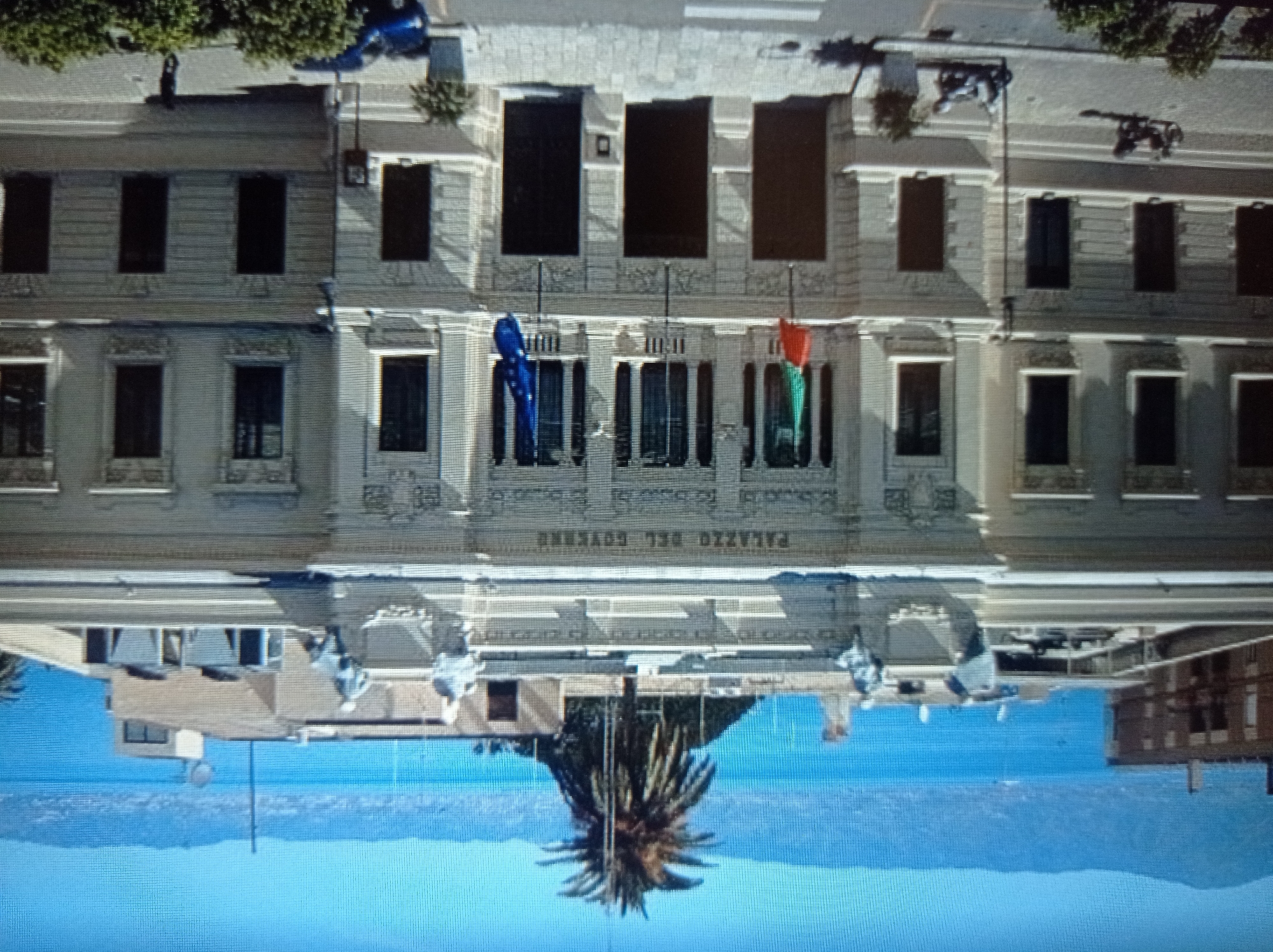 The image shows a large, ornate building, likely a government building, with the inscription "PALAZZO DEL GOVERNO" above its central entrance. The building is light-colored, possibly stone or stucco, and features classical architectural elements like columns, detailed carvings, and multiple windows. Two flags, one Italian and one European Union flag, are visible on poles in front of the building.
In front of the building, there are several elements:
- A street with visible white lines, suggesting a crosswalk.
- Two parked motorcycles, one dark-colored and one lighter-colored.
- A parked blue car.
- Potted plants are placed near the building's entrance and on the sidewalk.
- In the background, a body of water (likely a sea or large lake) is visible, with mountains or hills in the distance. A prominent palm tree stands on a rooftop to the left of the main building.
There are no people clearly visible in the image, though a shadowy figure can be seen near the blue car. The overall impression is a sunny day in a coastal or lakeside city with significant architectural heritage.
This image is NOT located in Mukachevo, Ukraine. The inscription "PALAZZO DEL GOVERNO" indicates an Italian-speaking location, and the Italian flag further supports this.
