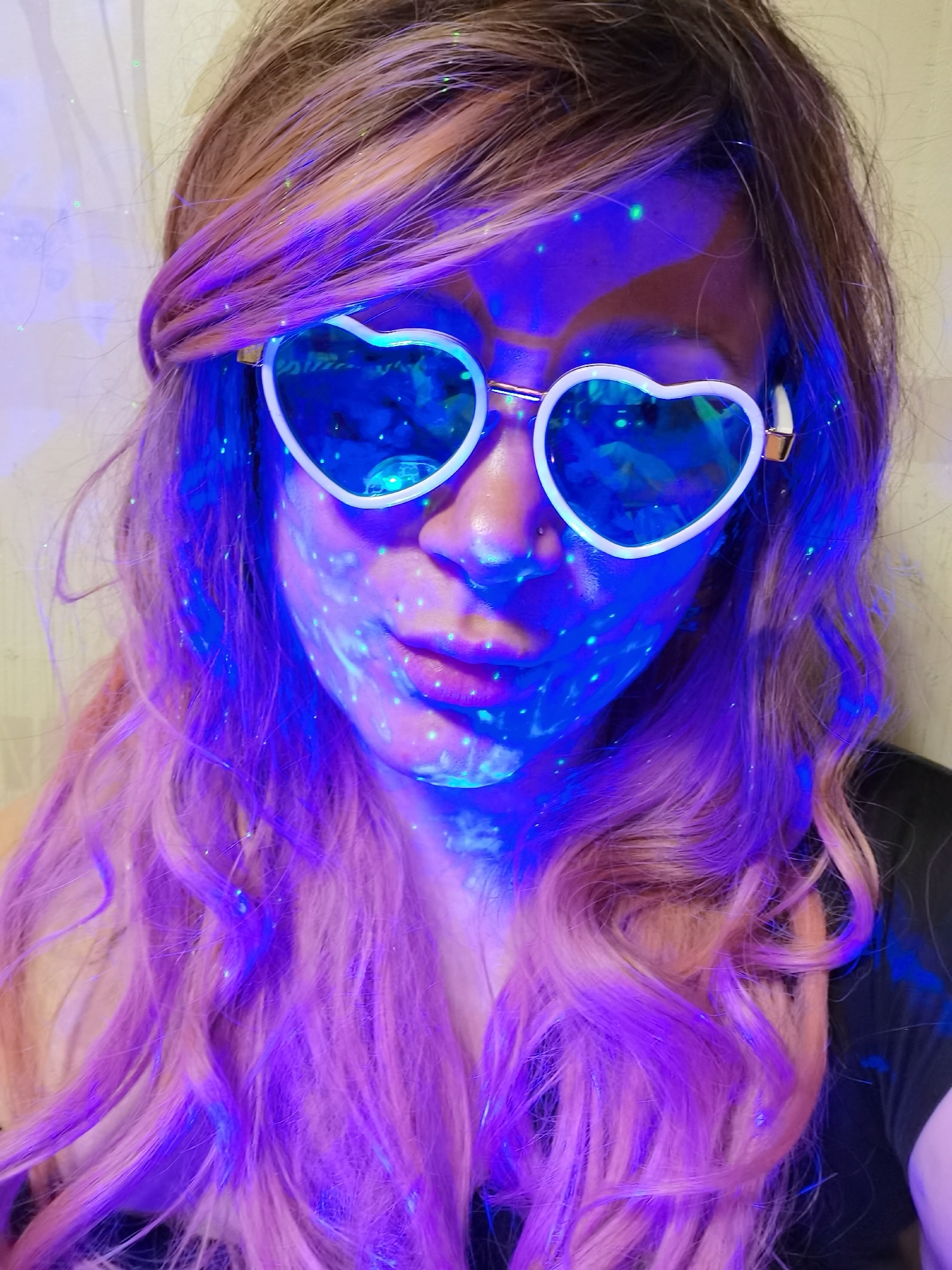 A close-up of a person with fair skin and pink hair, possibly a woman. She is wearing white-rimmed heart-shaped sunglasses with blue lenses. Her face and hair are illuminated with blue and green light, creating scattered dot patterns on her skin. The setting is indoors, against a plain, light-colored wall.
There is no indication that this image was taken in the city of Chaikovsky, Russia. The background and subject matter offer no specific geographic clues.