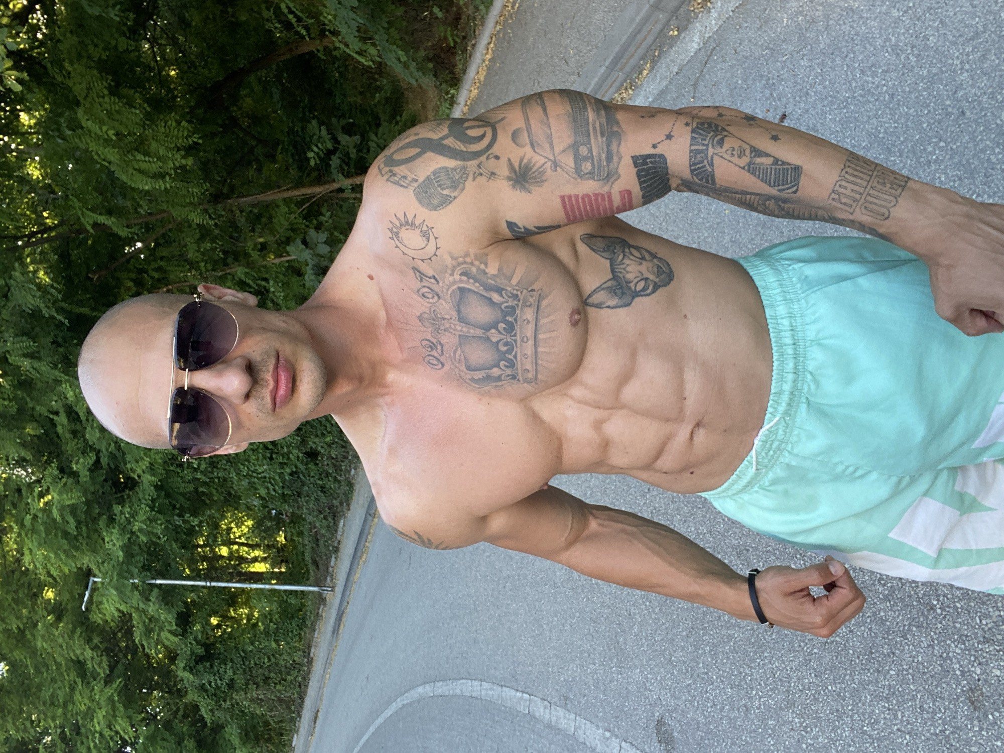 A bald man with sunglasses and an athletic build is standing on a paved road next to a grassy embankment and trees. He is shirtless, wearing light blue shorts. His chest and arms are covered in tattoos. The road curves to the left and appears to be in a park or a less populated area. The lighting suggests it is daytime, possibly late afternoon due to the angle of the sun. No specific landmarks indicating the city of Pleven, Bulgaria, are visible in the image.