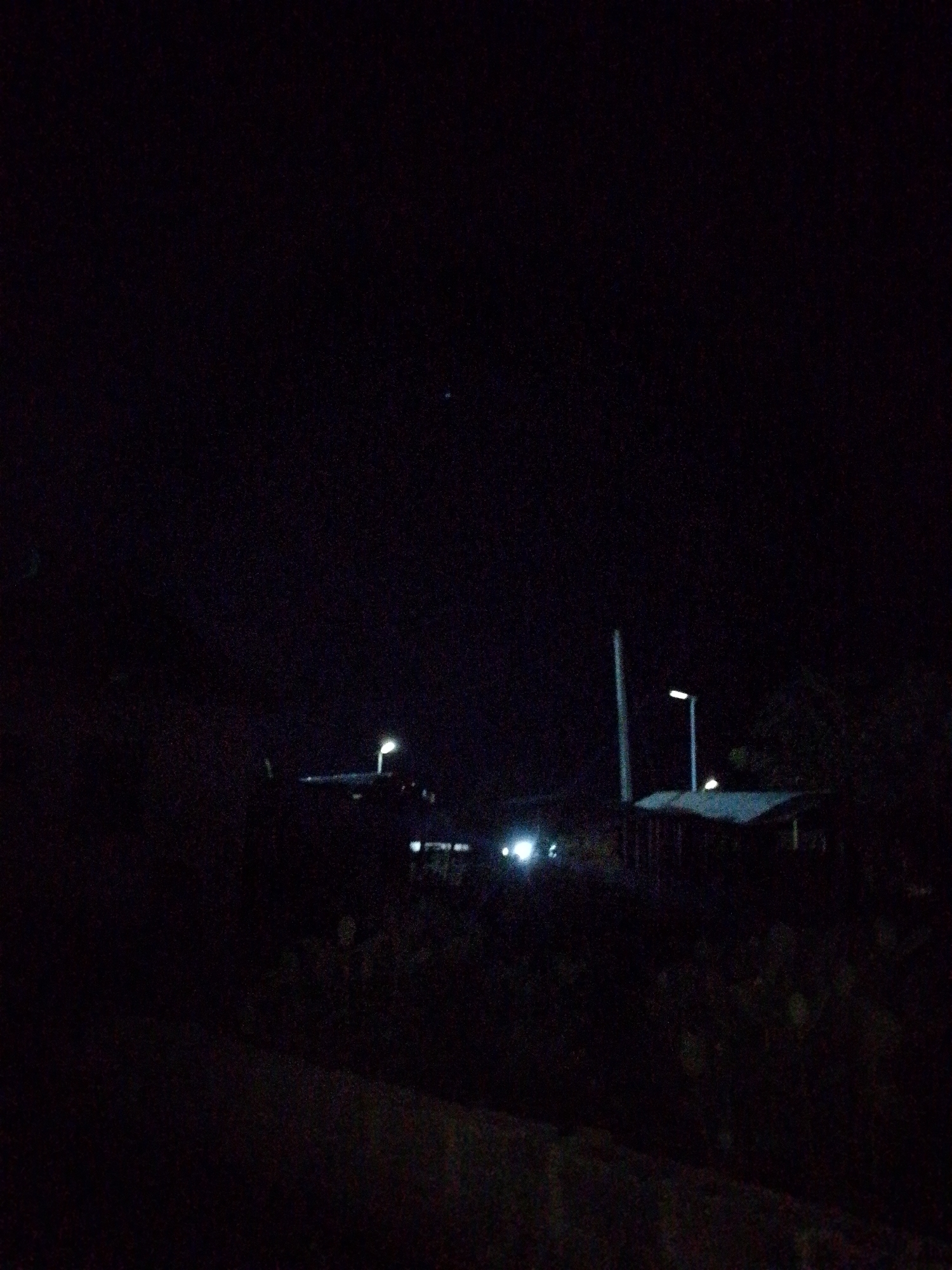 The image depicts a night scene in Adankolo, Nigeria. The scene is poorly lit, and visibility is low. There appear to be a few structures or buildings, likely residential or commercial, illuminated by distant streetlights. The presence of vehicle headlights suggests a road or path. The environment appears to be a mixture of vegetation and built infrastructure. No human figures are clearly visible.