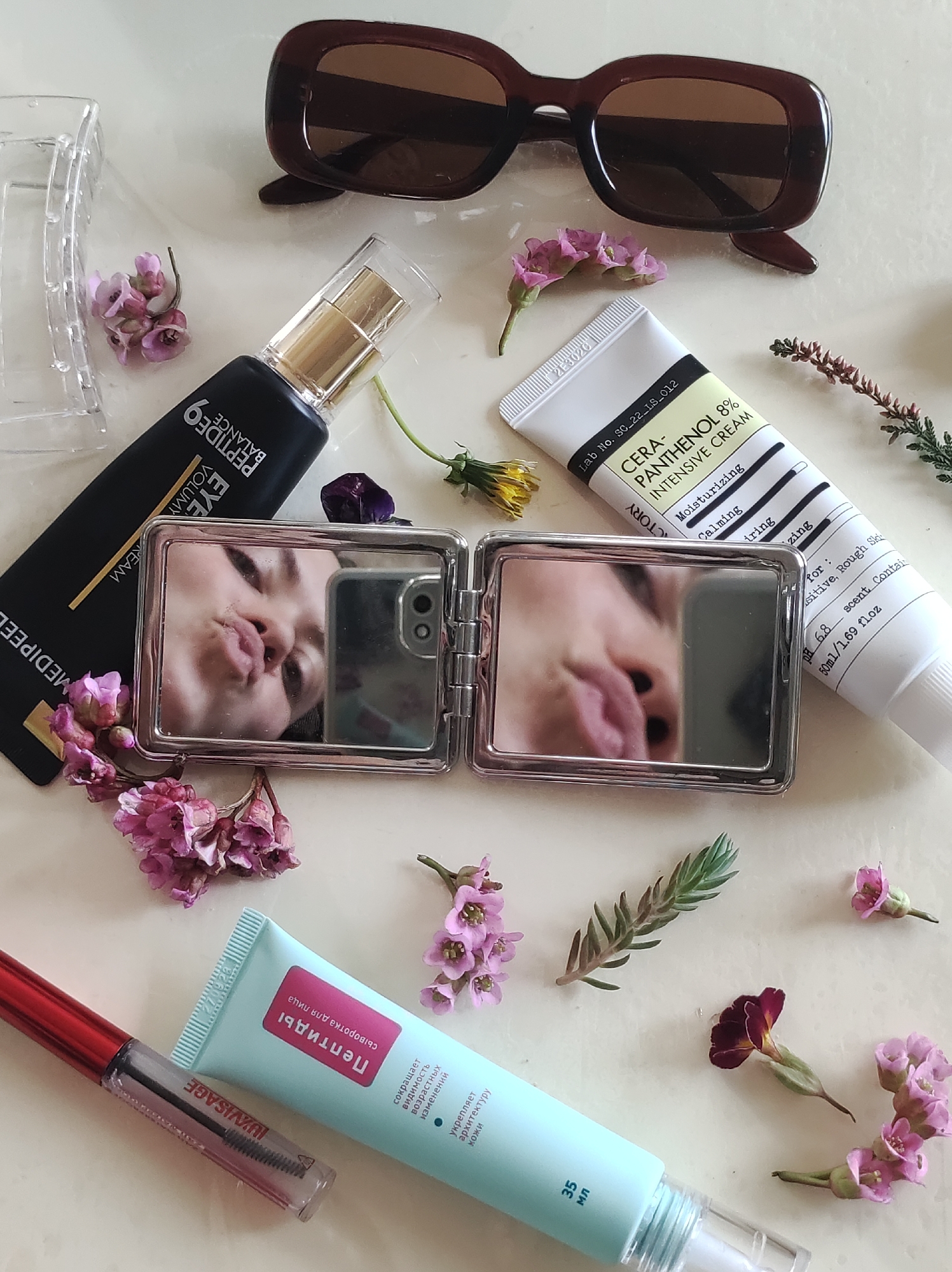 The image shows a flat lay of beauty products and accessories arranged on a white surface. At the top is a pair of brown sunglasses and a clear plastic hair clip. A black bottle labeled "MEDIPEEL Peptide 9 Volume Eye Cream" with a gold pump nozzle is positioned near a small, silver, folding mirror that reflects a person's face making a kissing expression and holding a phone to capture the reflection. To the right of the mirror is a white tube labeled "CERA-PANTHENOL 8% Intensive Cream". At the bottom, a light blue tube labeled with Russian text and "35 мл" is next to a clear mascara or brow gel tube with a red cap and "JUVISAGE" written on it. Scattered throughout the arrangement are sprigs and blooms of pink and purple flowers, and some greenery. The image does not suggest the location in the city of Фоки, Russia.