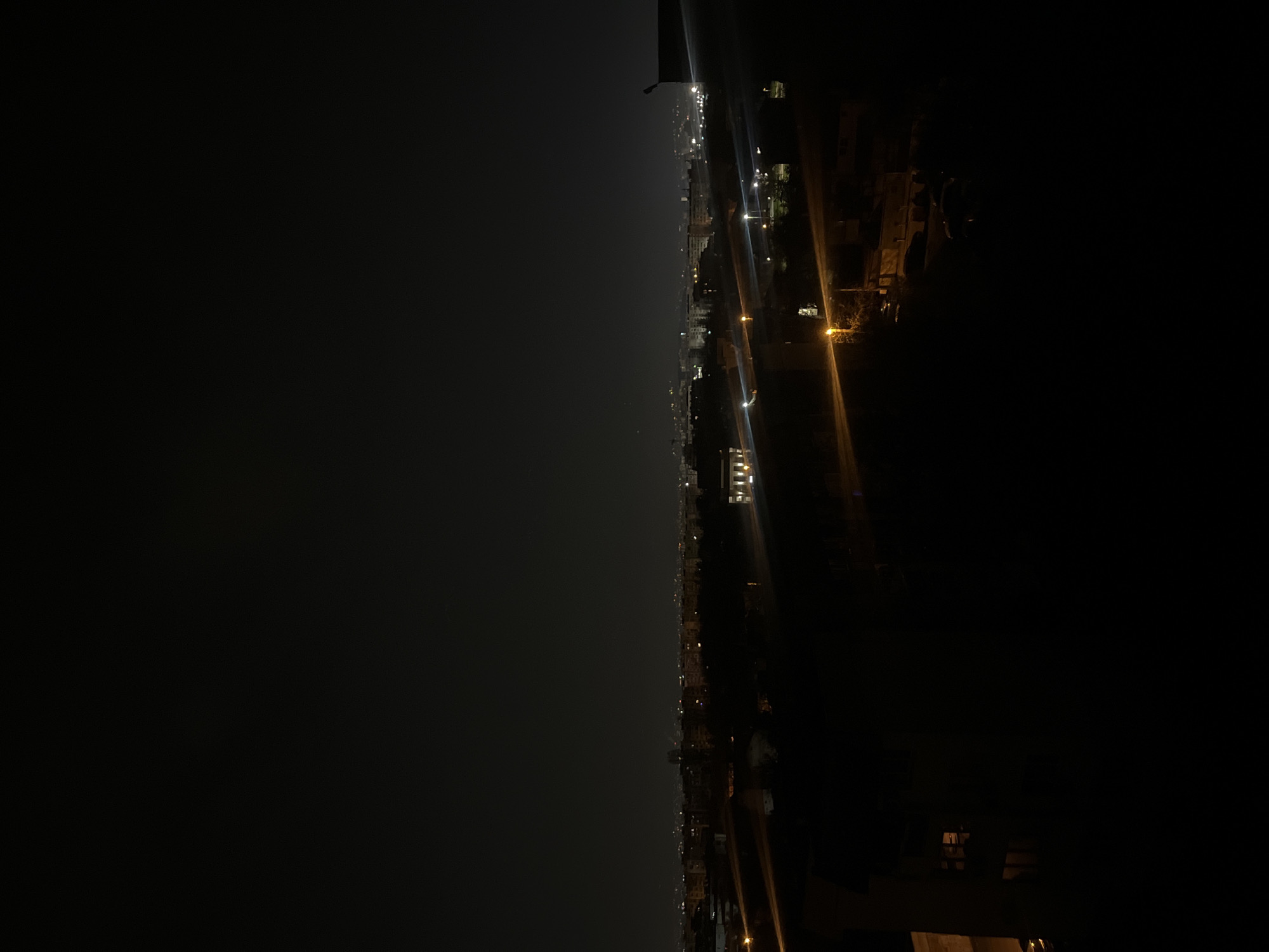 This image depicts a nighttime view of what appears to be a cityscape with numerous lights, taken from an elevated position, possibly a high-rise building or hill. The left side of the image is shrouded in darkness, likely indicating a clear, starless night or a lack of illumination. The right side shows a scattering of lights suggesting buildings, streets, and possibly vehicle traffic. In the distance, a denser concentration of lights suggests the city center or a more built-up area. The specific location in Sofia, Bulgaria, cannot be determined from this image alone. There are no discernible people, actions, or interactions visible.