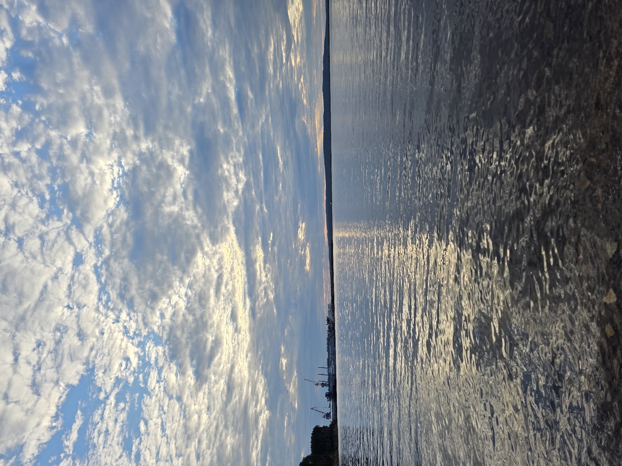 This is a landscape photograph taken from a low angle, showing a body of water and the sky. The horizon line is slightly tilted.
On the left side of the image, the sky is filled with scattered, fluffy white clouds against a blue sky. The sunlight catches the edges of the clouds, giving them a bright, luminous appearance. In the distance, along the horizon, there is a faint silhouette of what appears to be a coastline with some structures, possibly including cranes, suggesting an industrial or port area. The light on the horizon also hints at either sunrise or sunset.
On the right side of the image, the water is choppy and reflects the sky and light. The surface of the water is textured with ripples and waves, creating a dynamic pattern of light and shadow. The colors in the water are predominantly dark blues and grays, with streaks of bright light from the reflection. The rightmost part of the image shows a darker, more textured area, possibly the edge of the water or a rocky embankment.
The overall impression is one of a serene natural scene with hints of human activity in the distance. The time of day appears to be either early morning or late evening, given the quality of light. The weather seems clear but with some cloud cover. The location context suggests this could be a waterfront area in Chaikovsky, Russia, possibly near a port or industrial zone. There is no visible text in the image.