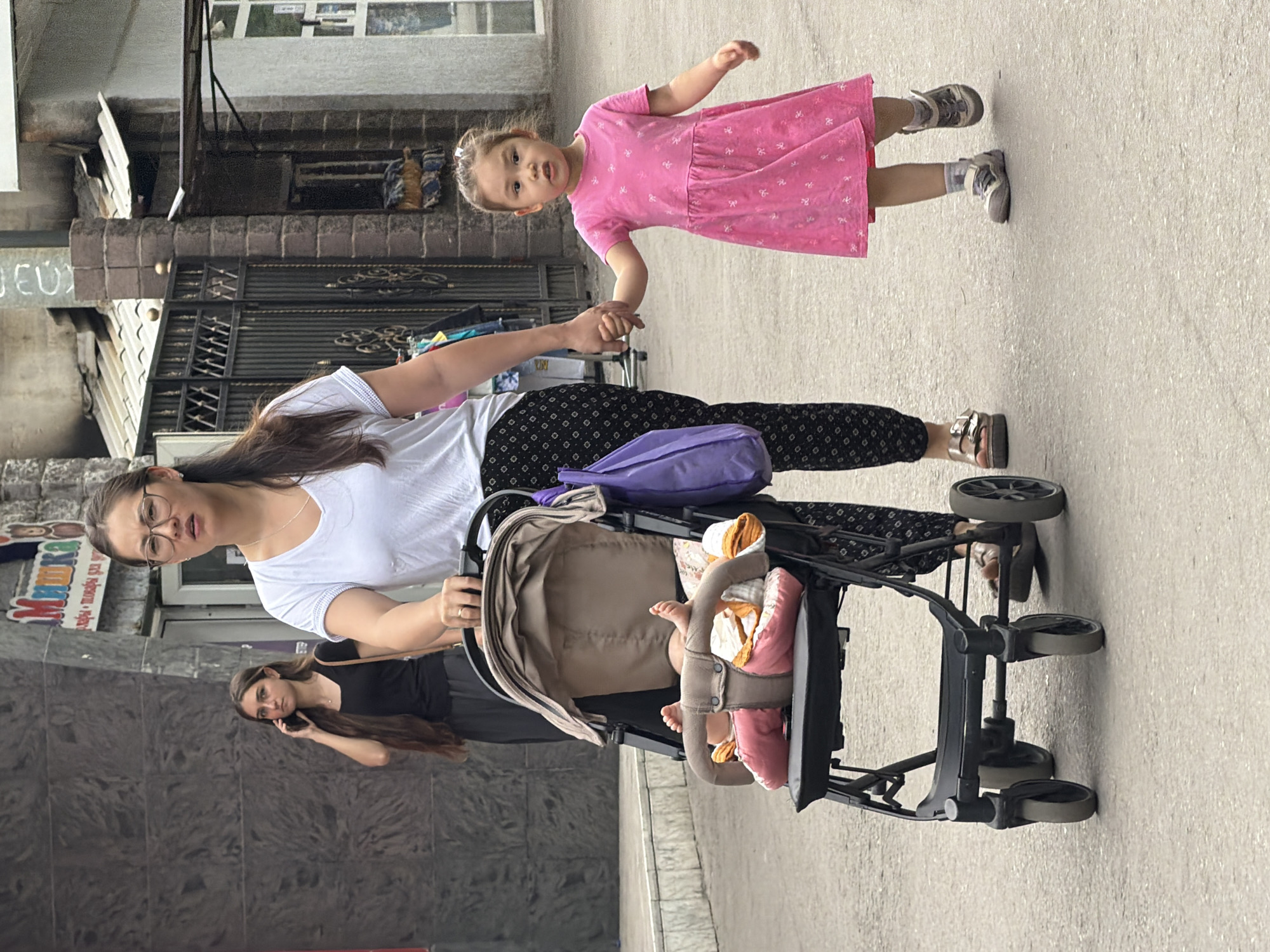 A woman wearing a white t-shirt and black patterned pants pushes a stroller with a baby inside, while holding hands with a young girl in a pink dress. The woman wears glasses and has her hair pulled back. The baby in the stroller appears to be wearing pink pants and an orange and white blanket. In the background, another woman with long brown hair is visible talking on a cell phone. The scene takes place on a paved street with buildings and a fence in the background.