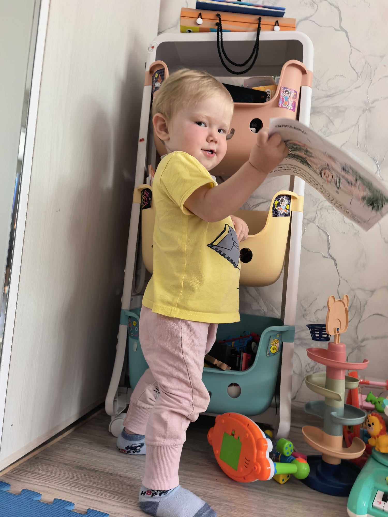 A blonde toddler, wearing a yellow t-shirt and pink pants, stands in a room and holds a book. The toddler is on the left side of the image, facing slightly to the right and looking at the camera. The toddler's left arm is extended, holding the book open. Behind the toddler is a white and pink shelf unit filled with toys. To the right of the shelf is a colorful toy ball ramp. The floor is wooden and a blue puzzle mat is visible in the lower left corner. The wall behind the shelf has a marble pattern. There is no discernible information in the image to determine its location in Sterlitamak, Russia.
