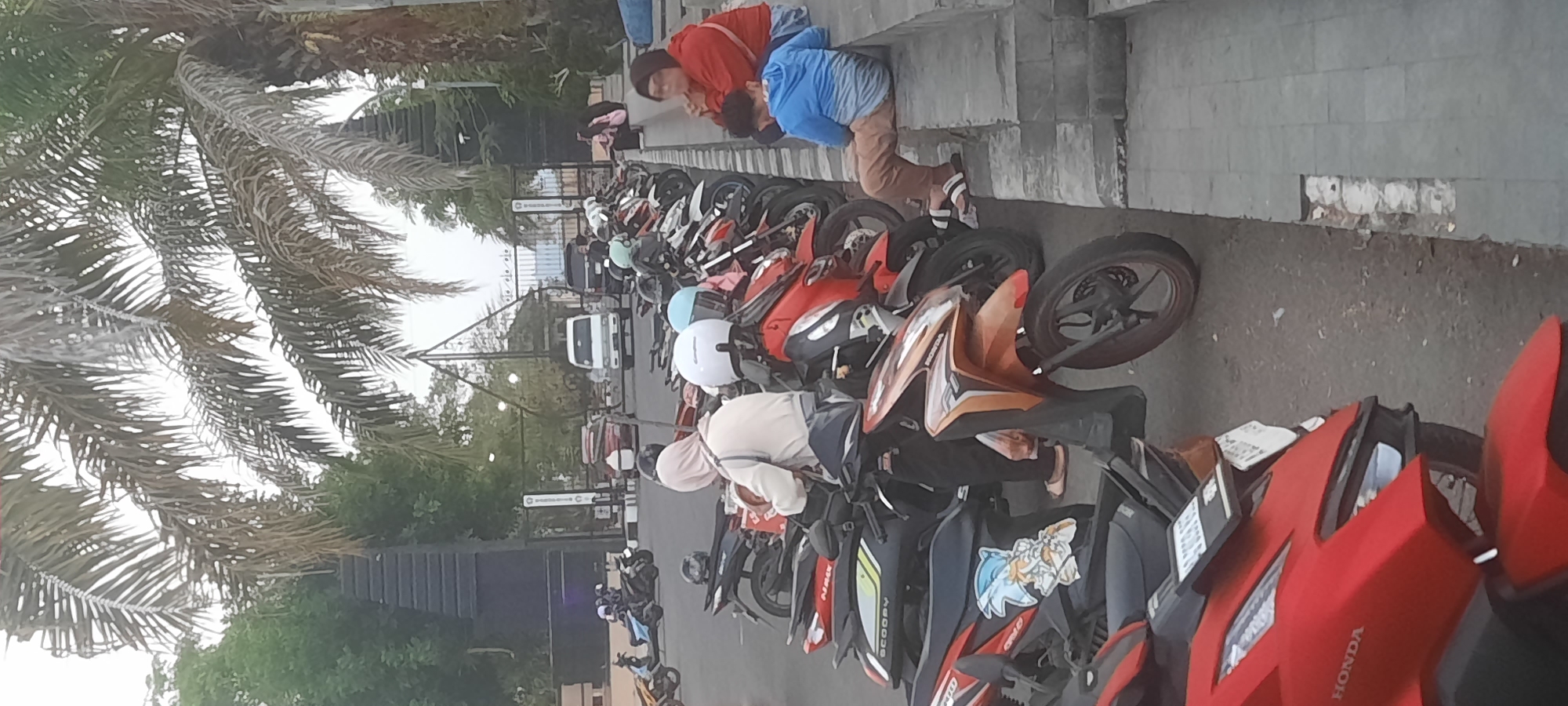 The image depicts a street scene in Kanigoro, Indonesia, featuring a row of parked motorcycles and several people. In the foreground, a red Honda motorcycle is visible, with a black and yellow scooter parked next to it, adorned with a cartoon sticker of Sonic the Hedgehog. Further down the row, more motorcycles are parked, including red and orange models.
Several individuals are present in the scene. A woman wearing a beige hijab and white top is standing next to a motorcycle, holding a red bag. To her right, a person in a blue hoodie and brown pants sits on a concrete ledge, facing away from the camera. Another woman, wearing a red jacket and dark headscarf, sits on the same ledge, looking down. In the background, a white van and a black SUV are visible, along with a dark gate and palm trees. The environment appears to be an outdoor area, likely a parking lot or street in a public space.