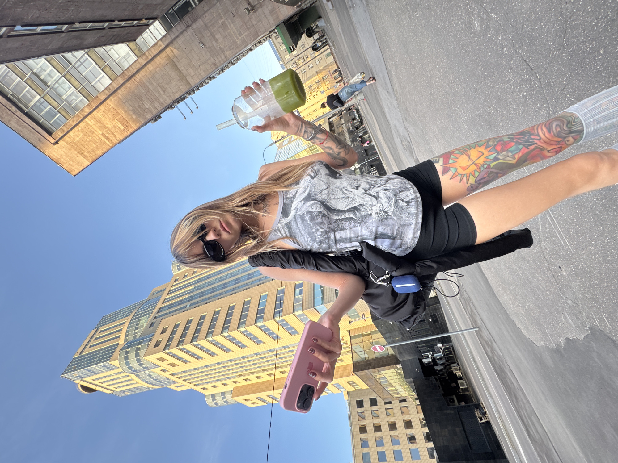 A young Caucasian woman stands on an asphalt street in Moscow, Russia, in front of a modern glass and concrete skyscraper. She is wearing sunglasses, a gray tank top with a graphic print, black shorts, and tall gray socks. Her right leg is extensively tattooed with colorful images, including a sun and abstract figures. She is holding a pink smartphone in her left hand and a clear plastic cup with a green beverage and straw in her right hand. A black backpack with a blue earbud case attached hangs from her left shoulder. The background shows the skyscraper, other buildings, a clear blue sky, and a few other pedestrians walking in the distance. The image is taken from a low angle, emphasizing the height of the buildings.
