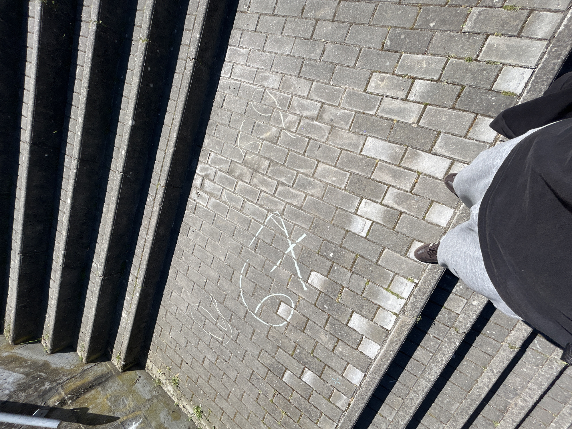 The image depicts an overhead view of a set of concrete steps and a paved area, likely in an outdoor urban environment. A person is standing on the paved area near the edge of the steps, wearing grey sweatpants, brown shoes, and a black jacket. White chalk drawings, including an "X", and an arrow can be seen on the paved area. The location appears to be in Mirandela, Portugal, based on the architectural style of the steps and paving.