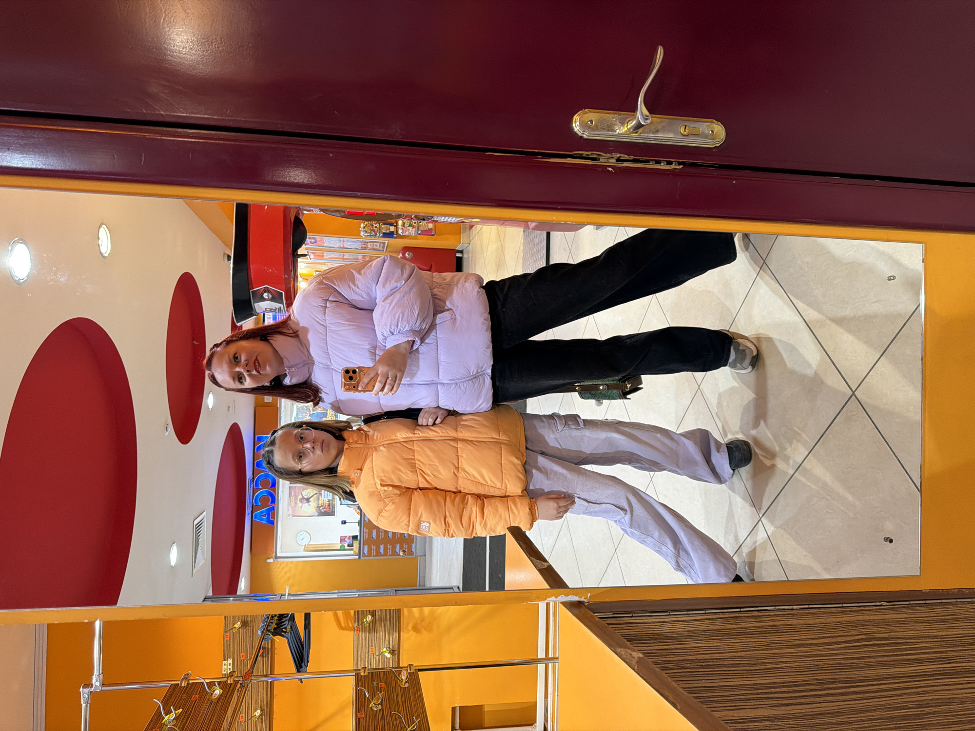 Two females are reflected in a mirror within what appears to be an indoor shopping environment. The upper female wears a light purple jacket and black pants, holding a phone in her hand. The lower female wears an orange jacket and light grey pants. The mirror has a red frame at the top and an orange frame at the bottom. The location appears to be a shop, evident from the clothing racks and retail displays reflected in the mirror. The background displays a red and white ceiling design. The signage appears to read "ACCA," suggesting a possible retail store location. There's no definitive element in the image that indicates Chaikovsky, Russia, specifically.