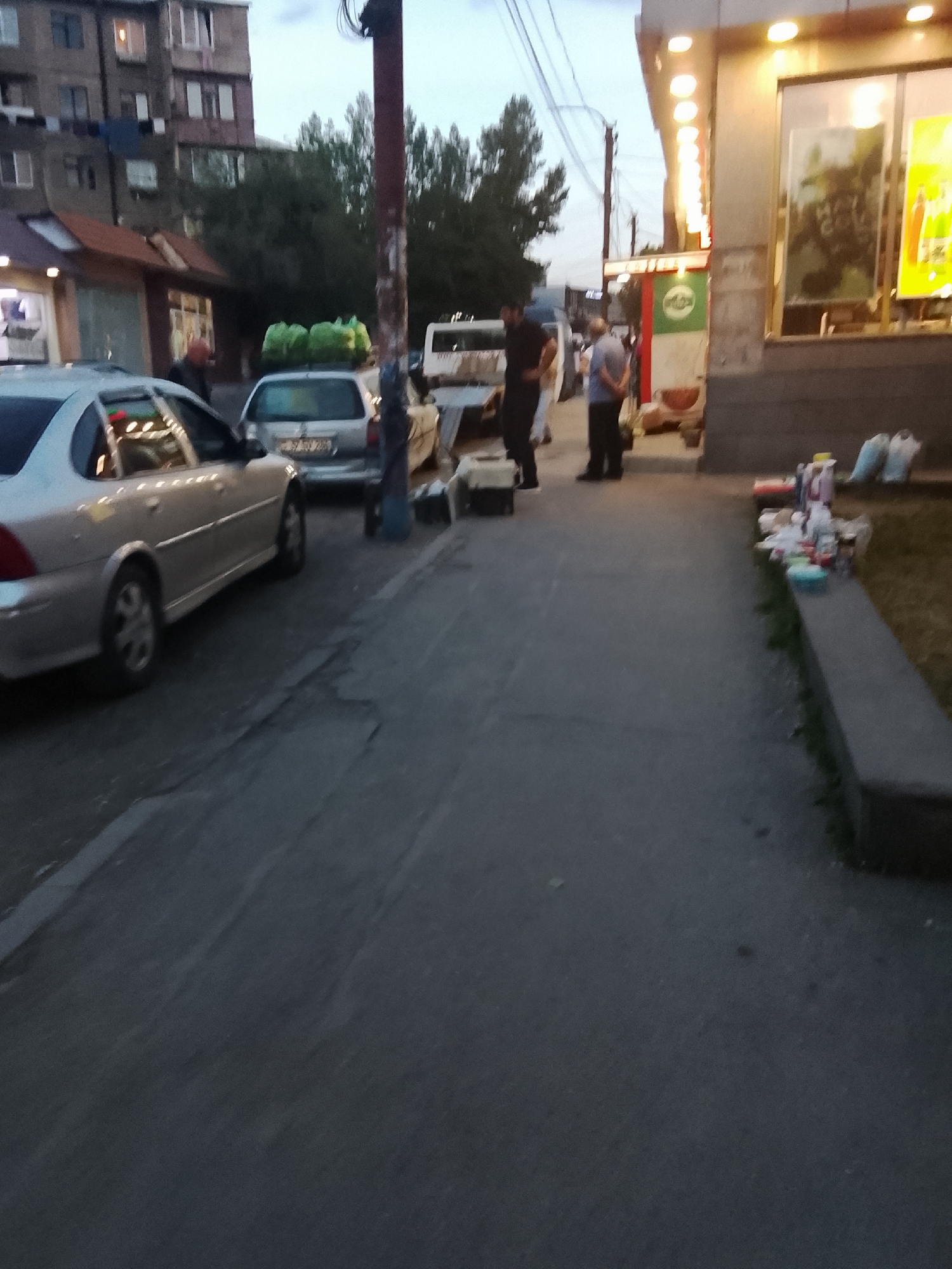 In an outdoor urban setting at dusk, a silver sedan is parked on the left side of a paved area. Behind it, a silver hatchback is also parked. Several people are standing on the sidewalk to the right. One individual in a dark shirt and pants stands near the hatchback. Another person in a light-colored shirt and dark pants is further right, facing away from the camera. A third person, also in dark pants and a light shirt, stands next to them. A store with a lit facade is visible on the right, with various items displayed on a ledge in front of it. Trees are present in the background. The sky is overcast, suggesting evening.