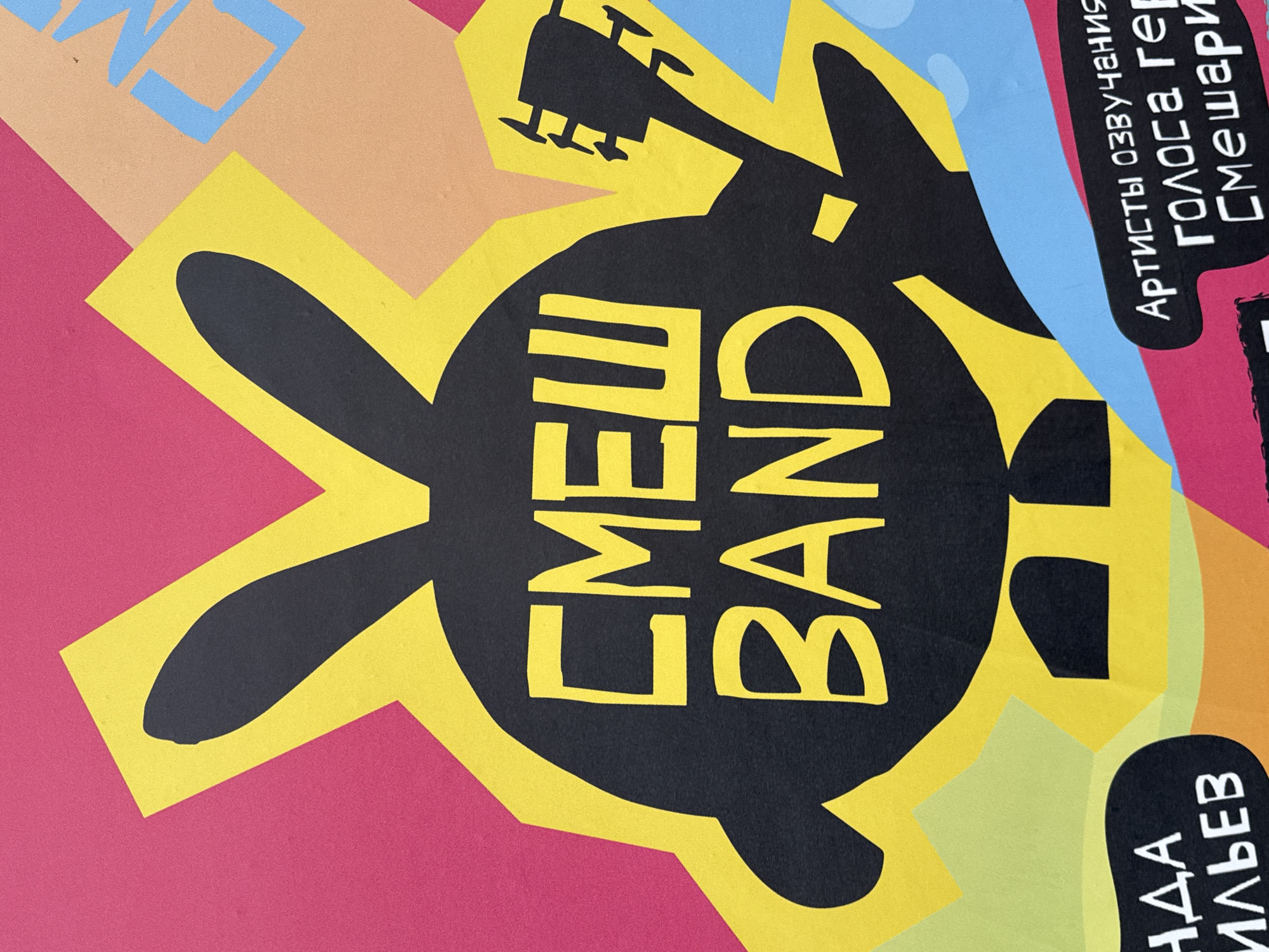 The image shows a poster or advertisement in Saint Petersburg, Russia. It features a black silhouette of a rabbit head with a guitar superimposed over it. The text "CMEW BAND" is written in yellow across the rabbit's body. Additional text in Russian, including "АРТИСТЫ ОЗВУЧАНИЯ ГОЛОСА ГЕР СМЕШАРИКИ," is present on a rectangular patch. The background is a collage of colorful geometric shapes in pink, tan, light blue, and yellow. Another fragment of text, "ИЛЬЕВ" is partially visible on a green background.