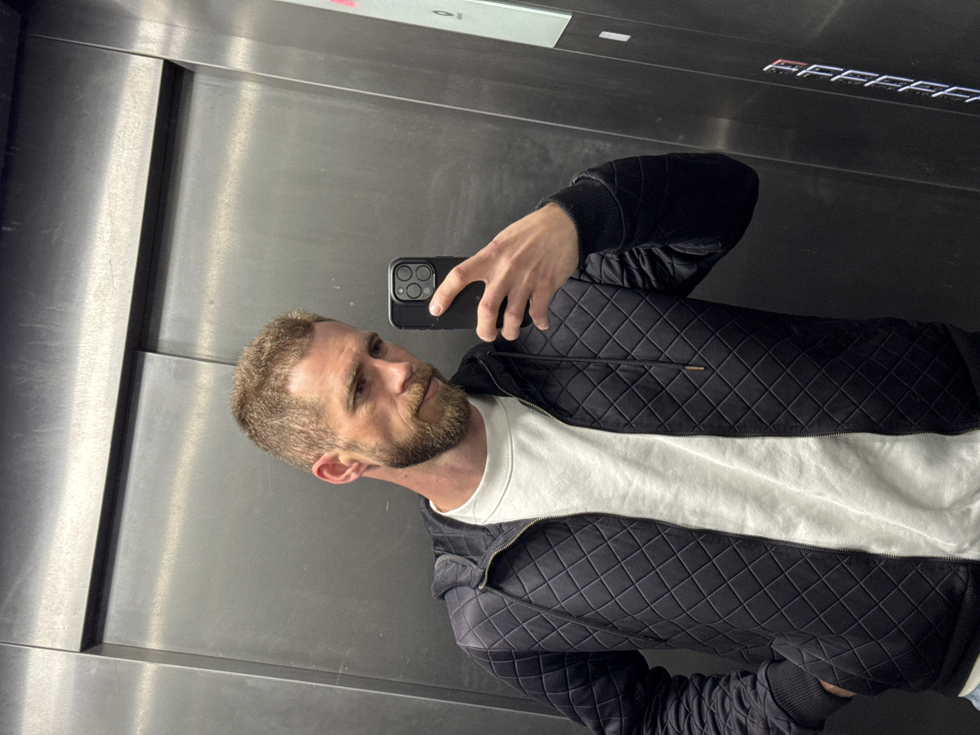 An adult male is captured in a mirror selfie taken inside a modern elevator, located in Budva, Montenegro. The image is oriented vertically, showing the man from approximately the chest up.
The man has short, light brown or blond hair and a neatly trimmed beard of the same color. He wears a black quilted jacket with a zipper, over a white, long-sleeved crew-neck shirt. His right hand holds a black smartphone, likely an iPhone given its distinct triple-lens camera module, positioned to capture his reflection. His gaze is directed slightly away from the phone, towards his own reflection, with a neutral and somewhat contemplative expression.
The elevator interior features brushed metallic walls, likely stainless steel, which reflect light and create subtle gradients. Above the man's head, an elevator control panel is visible, displaying blurry white and red digital text or numbers. On the upper right side of the image, appearing on the elevator's ceiling or a high panel, reflective silver characters spell out "FFFFFF". The overall setting is contemporary and indoors.