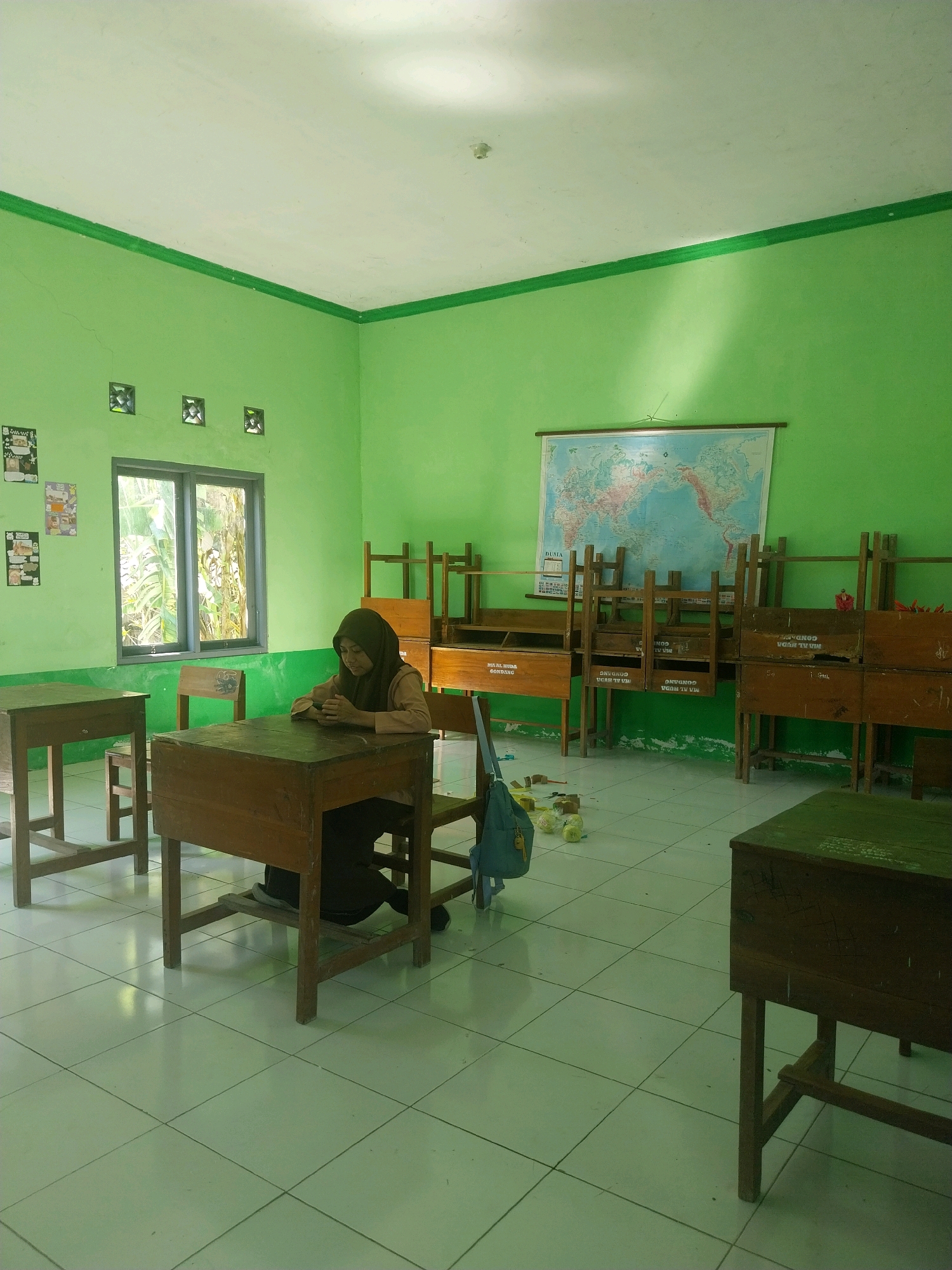 A young woman wearing a brown hijab and a tan top sits at a wooden desk in a classroom. The room has bright green walls and a white ceiling. A world map is displayed on the wall behind her. There are several other wooden desks and chairs in the room, some stacked up. A blue backpack is hanging on a chair. The floor is tiled with light green squares. The scene appears to be in Pandean, Indonesia.
