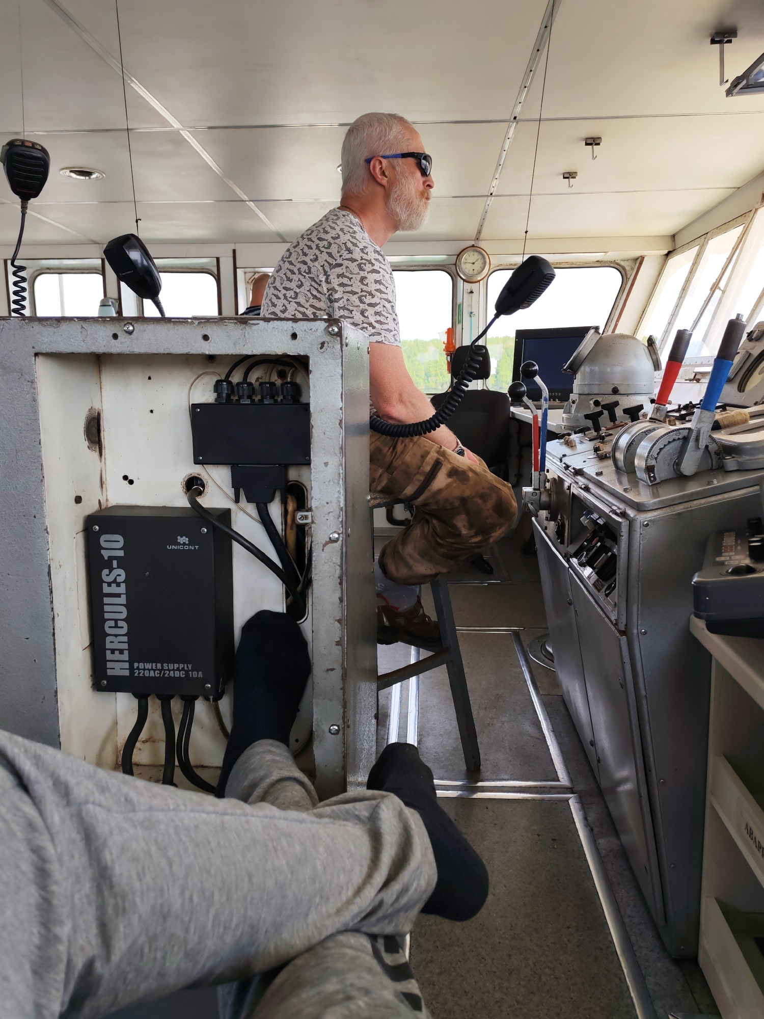 The image appears to be taken inside the wheelhouse of a vessel, possibly a boat or ship. One person, an older Caucasian male with a white beard and sunglasses, is seated on a stool and is turned to his right, looking out of the window. He is wearing a patterned shirt and brown pants. In the foreground, there are two legs clad in gray sweatpants, with black socks on the feet, that are extended toward the viewer, seemingly resting. There is an electrical box on the left of the image with the brand "UNICONT HERCULES-10" and the text "POWER SUPPLY 220AC/24DC 10A" on it. To the right of the seated male, there's a large control panel with various levers and switches. Microphones are hanging from the ceiling.
The location cannot be precisely determined as being in Khoroshevo, Russia based on the information in the image. There are no definitive visual elements in the photograph that specifically establish the city as Khoroshevo.