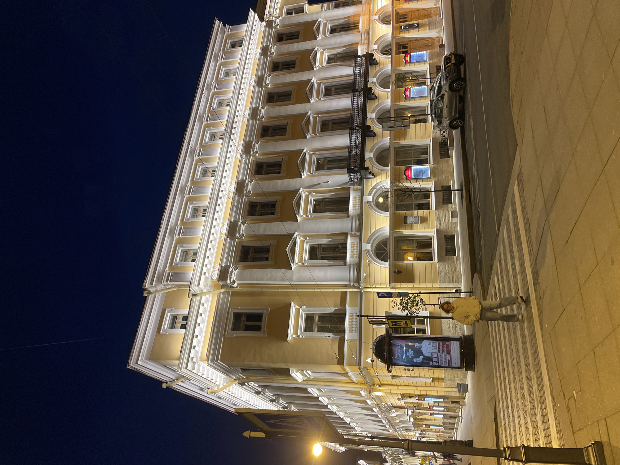 This night-time image captures a grand, multi-story building in Saint Petersburg, Russia, illuminated against a dark blue sky. The building features a pale yellow facade with extensive white architectural detailing, including cornices, elaborate window frames, pilasters, and decorative elements. Numerous upward-pointing lights highlight the intricate details of the building's exterior.
At street level, a paved road runs alongside the building, with a silver car parked by the curb. A wide sidewalk, composed of patterned light-colored paving stones, parallels the street. One person, dressed in a light jacket and jeans, is visible standing on the sidewalk near a prominent cylindrical advertising pillar.
The scene is clearly set at night, with streetlights and the building's illumination providing the primary light sources. The sky is clear, with no visible clouds.
The advertising pillar displays text, including "24 часа" (24 hours) and "Музыканты" (Musicians), accompanied by images of people, suggesting an event or service. Other smaller signs on the building's lower levels have some visible Cyrillic text, but are mostly illegible. The overall atmosphere is calm, showcasing the architectural beauty of the city at night.