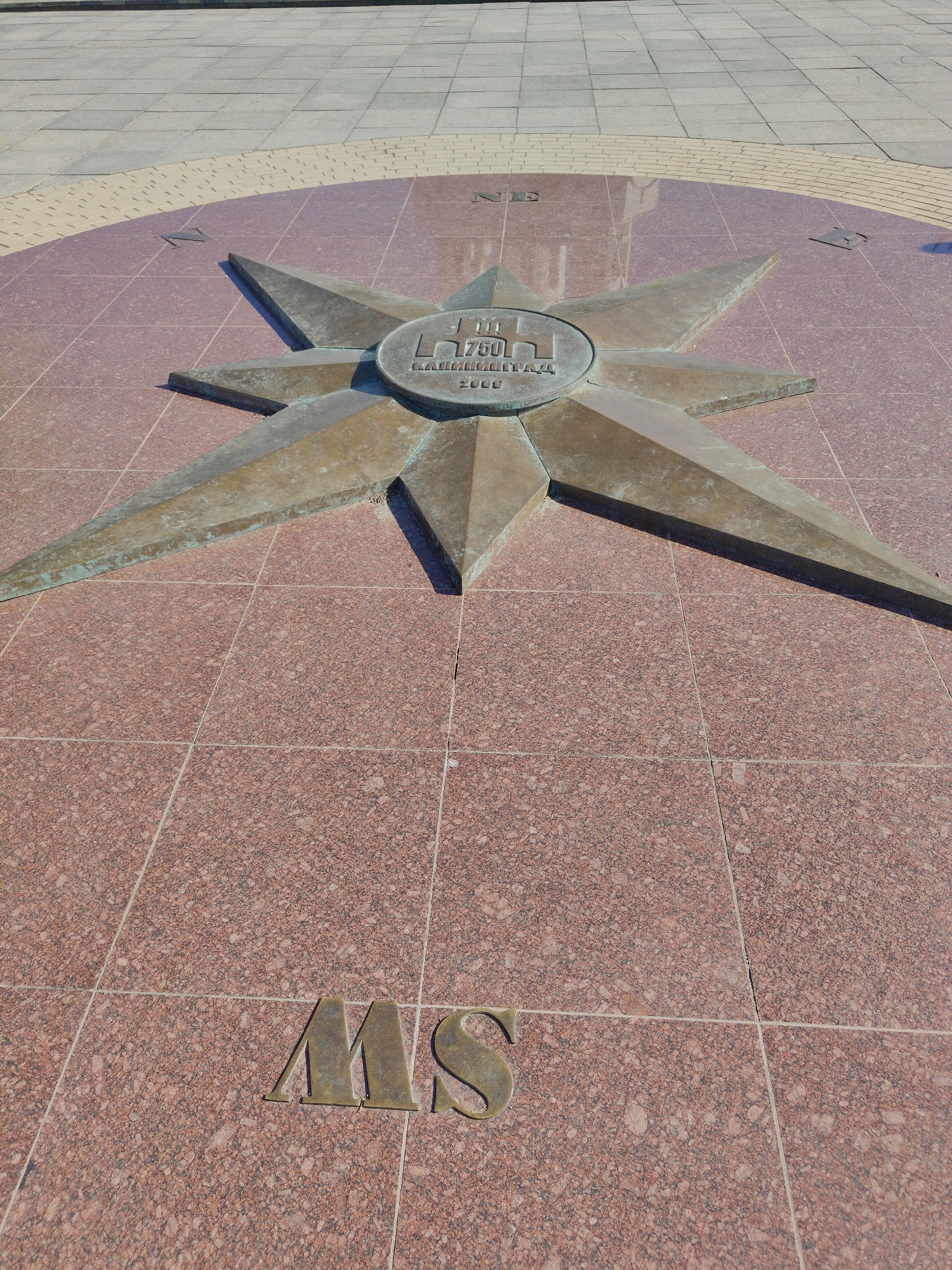 The image depicts a compass rose inlaid into a tiled surface. The compass rose is made of a tarnished metal, likely bronze or brass, and features a central emblem bearing the inscription "750 Kaliningrad 2006." The cardinal directions are marked with the letters "NE," "MS," and what appears to be "NW" and "SE," indicating northeast, southwest, northwest, and southeast respectively. The tiles surrounding the compass rose are a reddish-brown color with a speckled texture. The background shows a paved area with grey tiles, and a border of smaller beige tiles. The absence of any people or moving objects suggests a static, possibly public, location. The inscription implies a commemorative element, possibly marking the 750th anniversary of the city of Kaliningrad.