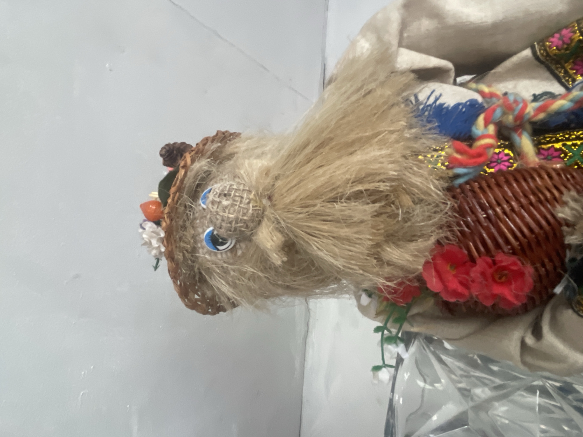 This is a close-up, eye-level, outdoor photograph of a straw doll with a long, tan beard and blue eyes. The doll is wearing a woven straw hat adorned with small white flowers, an orange berry, and a brown pinecone. The doll's clothing is visible, including a beige garment with colorful embroidered trim. A small woven basket filled with red flowers is attached to the doll's front. The background consists of a light-colored wall or surface. The image is undated and its location in Nikolayevskaya, Russia, is not discernible from the visual information provided.