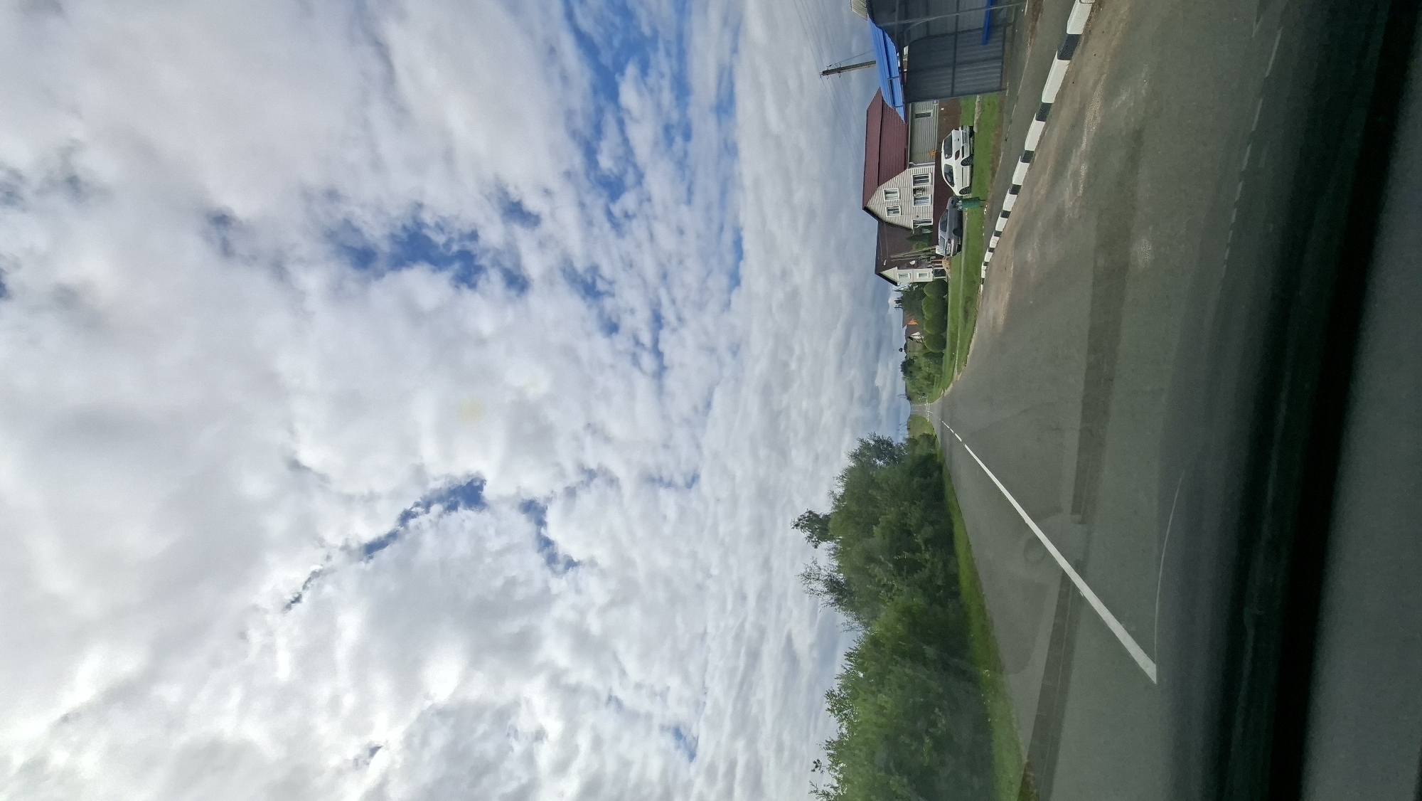 The image shows a view from a vehicle traveling on a paved road in a rural area. The road has a white dividing line. On the left side of the road is a dense line of green trees and bushes. On the right side of the road, there are houses, cars, and a bus stop. The sky is filled with white and grey clouds, with patches of blue sky visible. The scene appears to be in Myadzyel, Belarus, given the context. There are no people clearly visible in the image. The overall impression is a quiet, suburban or rural landscape on a cloudy day.