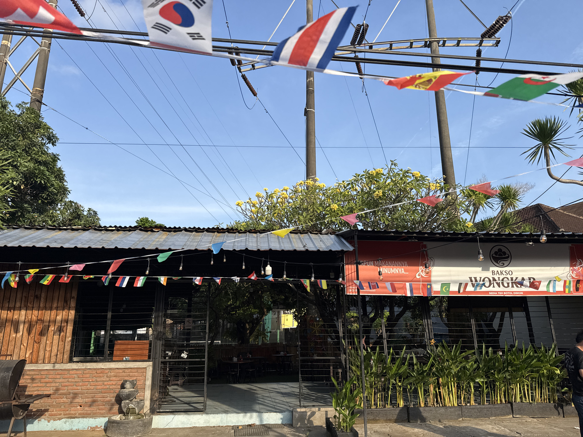 The image depicts the exterior of a restaurant called "BAKSO WONGKAR" located in Denpasar, Indonesia. The restaurant has a rustic facade with wooden paneling and a brick base. A corrugated metal roof extends over the dining area, which is visible through large metal-barred windows and an open gate. Strings of small flags representing various countries are hung across the front of the building, some fluttering in the breeze. Decorative lights are also strung along the roofline. A large banner above the entrance features the restaurant's name and a slogan in Indonesian. Lush green plants are arranged in planters in front of the building, and a small decorative fountain is situated near the entrance. The sky is clear and blue, suggesting a bright, sunny day. A person is partially visible on the right side of the frame, wearing a dark t-shirt and jeans. The scene is a typical street-level view of a local eatery in a tropical urban environment.
