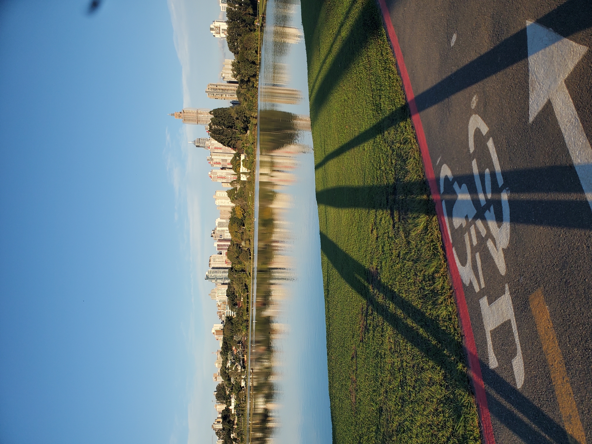 This is a photograph taken at the Parque Barigüi in Curitiba, Brazil, showing the lake with the city skyline reflected in it. In the foreground, a paved path with a bicycle symbol and an arrow is visible, bordered by a red line. Tall, modern buildings form the city skyline in the background, nestled among trees and green spaces. The sky is a clear blue. The sun is low, casting long shadows from unseen objects or people onto the grass and the path. The water of the lake is calm, creating a clear reflection of the sky and the cityscape. The overall scene depicts an urban park setting during a bright, sunny day.