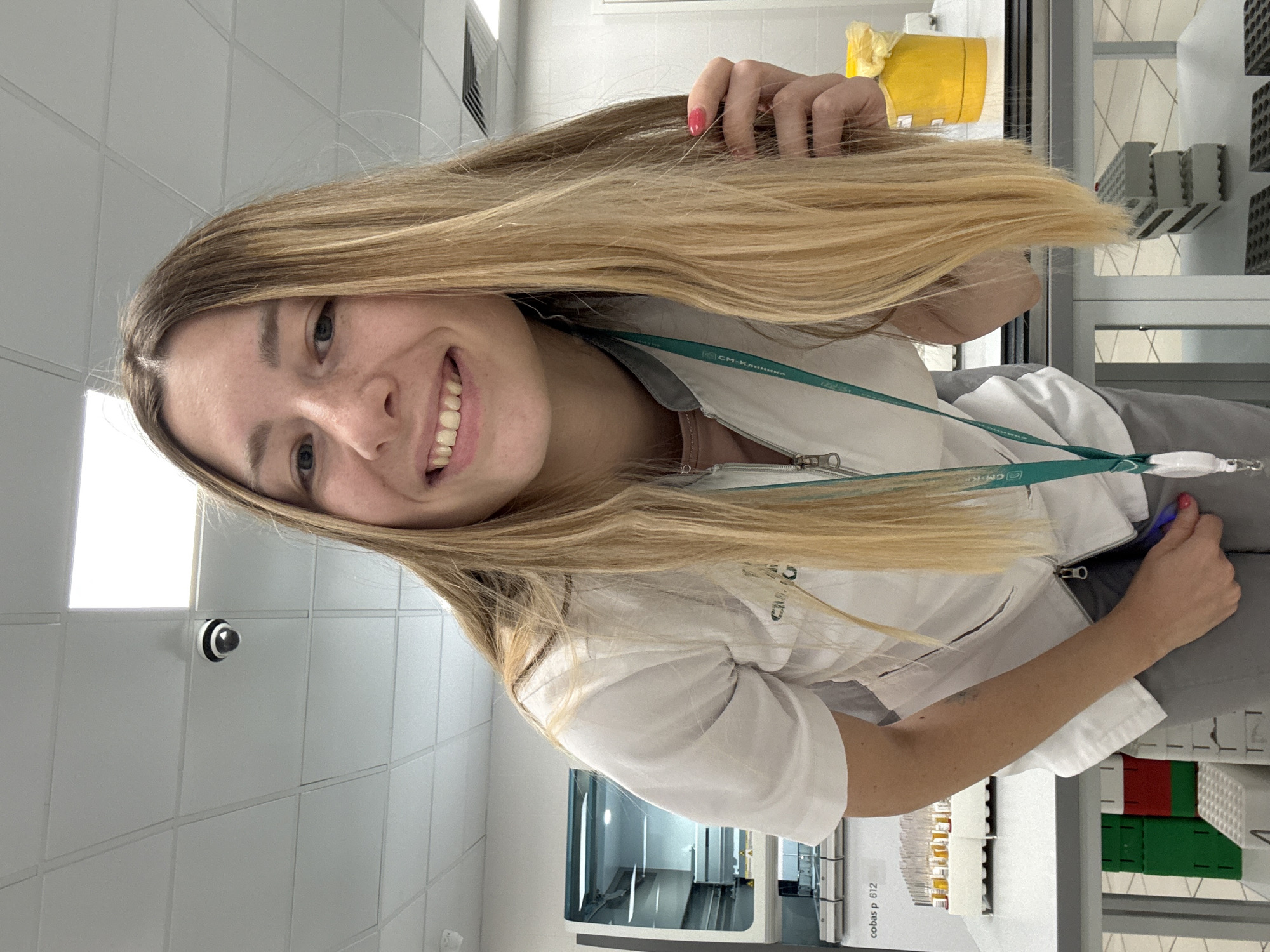 A young Caucasian woman with long, blonde hair, wearing a white lab coat over a pink shirt and grey pants, is leaning forward and smiling at the camera. She is in what appears to be a laboratory setting. To her left, a medical testing machine with a screen and various components is visible. In front of the machine, a rack of test tubes filled with yellow liquid is on a table. Her right hand is touching her hair, and her left hand is holding a blue object. A yellow bin is on a table to her right. The ceiling is made of white acoustic tiles with fluorescent lighting and a security camera is mounted on the ceiling. The overall environment suggests a clinical or research laboratory in Moscow, Russia.