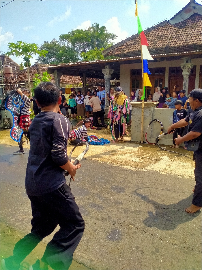 A group of people are gathered in front of a house in Kecamatan Kedawung, Indonesia (latitude: -7.52, longitude: 111.028). A man dressed in a colorful costume, a black and blue costume, and a man wearing a striped shirt are performing in the street in front of the house. The man in the striped shirt is lying on the ground, while the other two men are standing. In the background, a group of people are watching the performance. A man in black is holding a whip. The house is made of brick and has a tiled roof. There is a flagpole with a colorful flag attached to it. The flag is a Buddhist flag. There is a wooden gate to the left of the house. The street is paved with asphalt and has a dirt shoulder. The sky is clear and the sun is shining. There are trees and vegetation in the background.